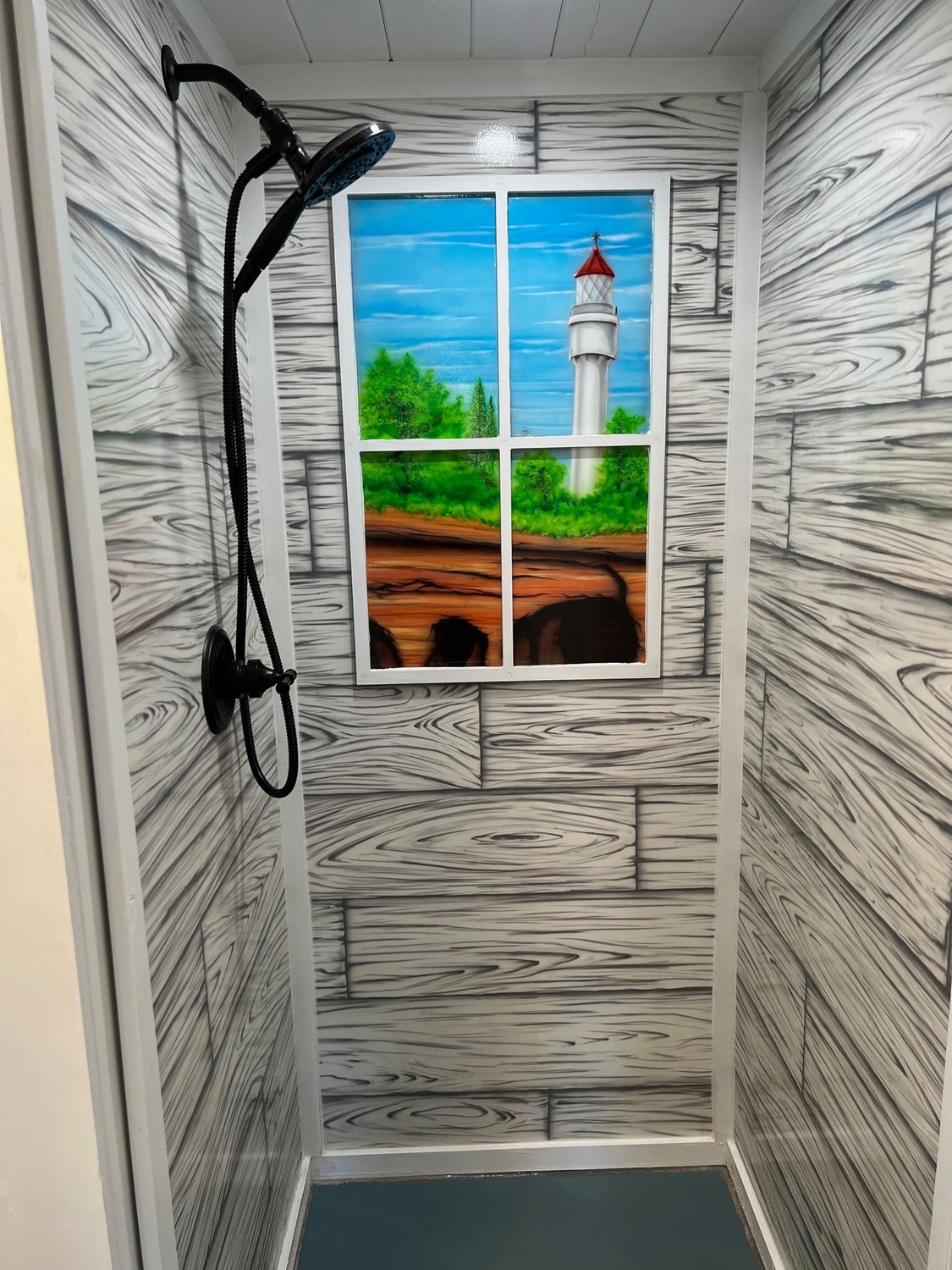 Shower stall with faux wood paneling, black showerhead, and window mural of a lighthouse.
