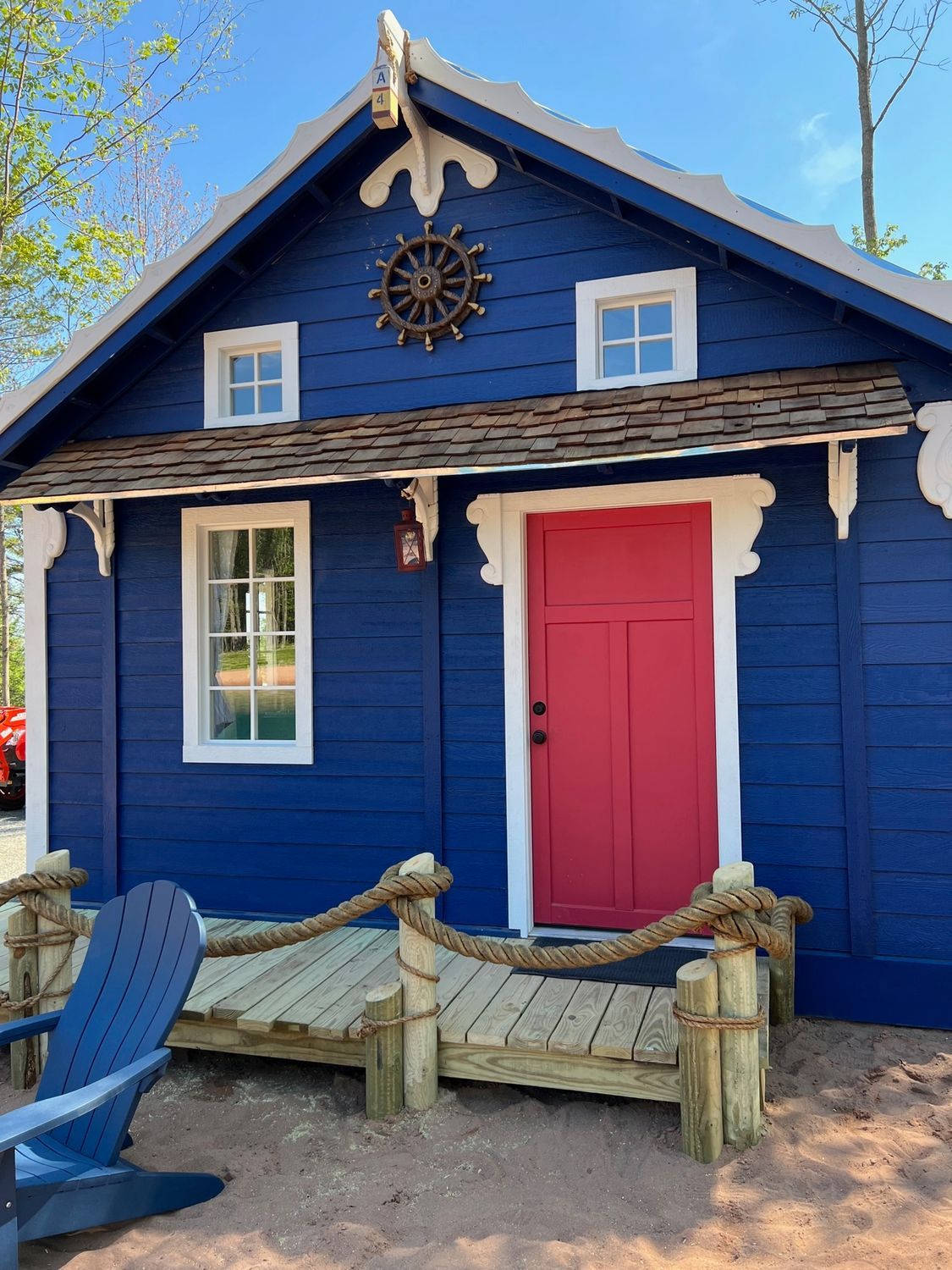 Blue nautical-themed cottage with red door, white trim, and wooden deck. Decorative ship wheel on the front.