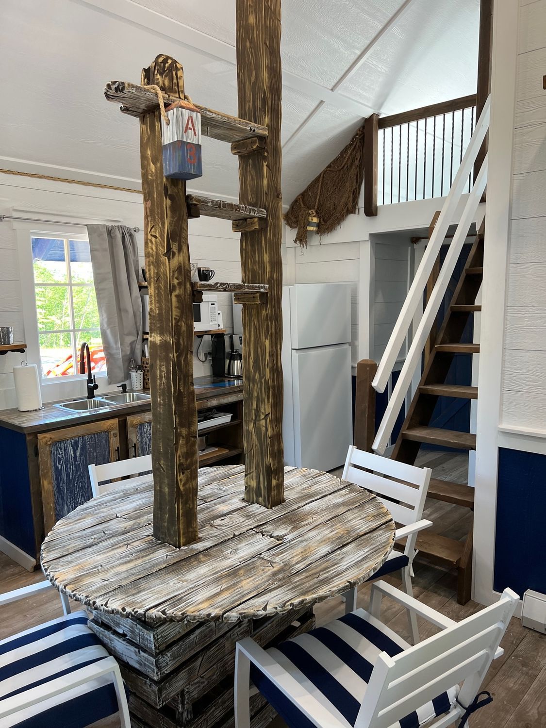 Rustic dining table with overhead wooden supports; blue and white striped chairs; kitchen area in the background.