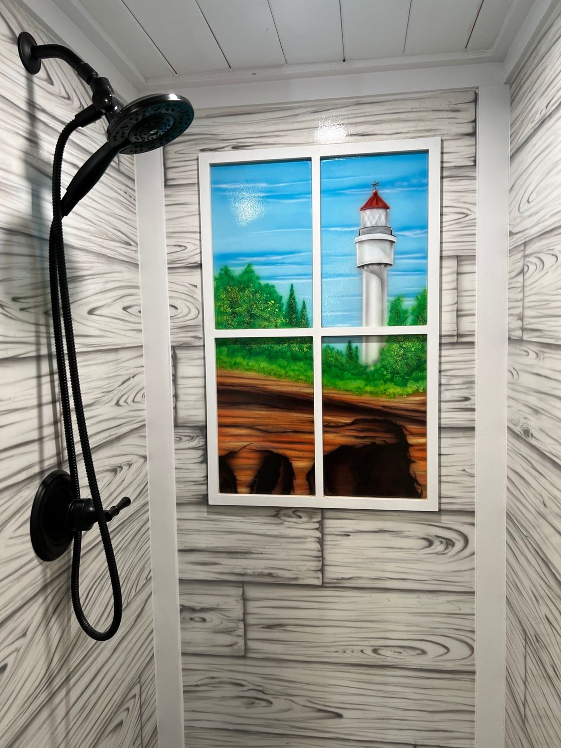 Shower with a black showerhead, faux wood paneling, and a painted window scene with a lighthouse.