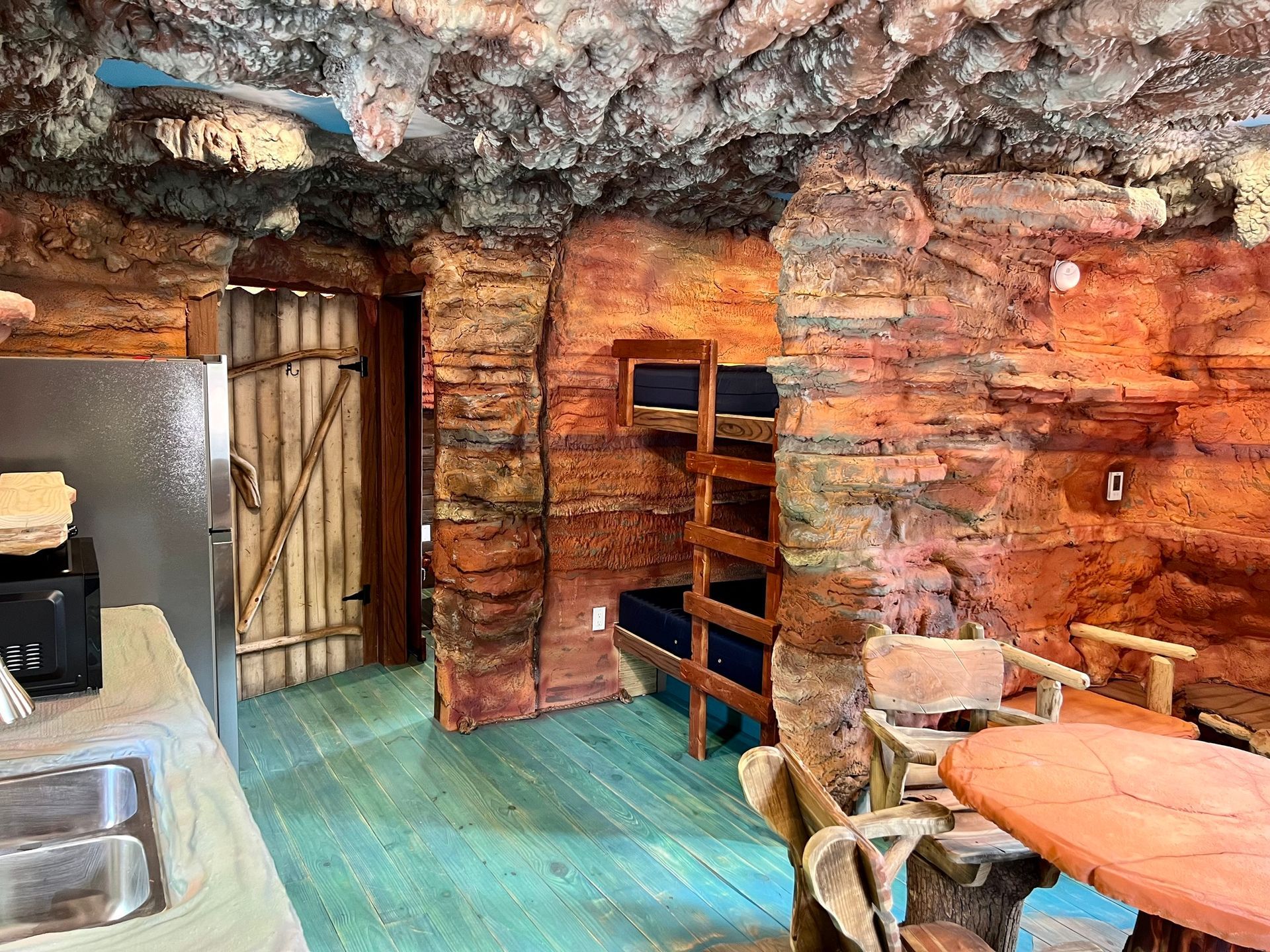 Interior of a cave-like room with reddish-brown walls, a bunk bed, a table, and a refrigerator.