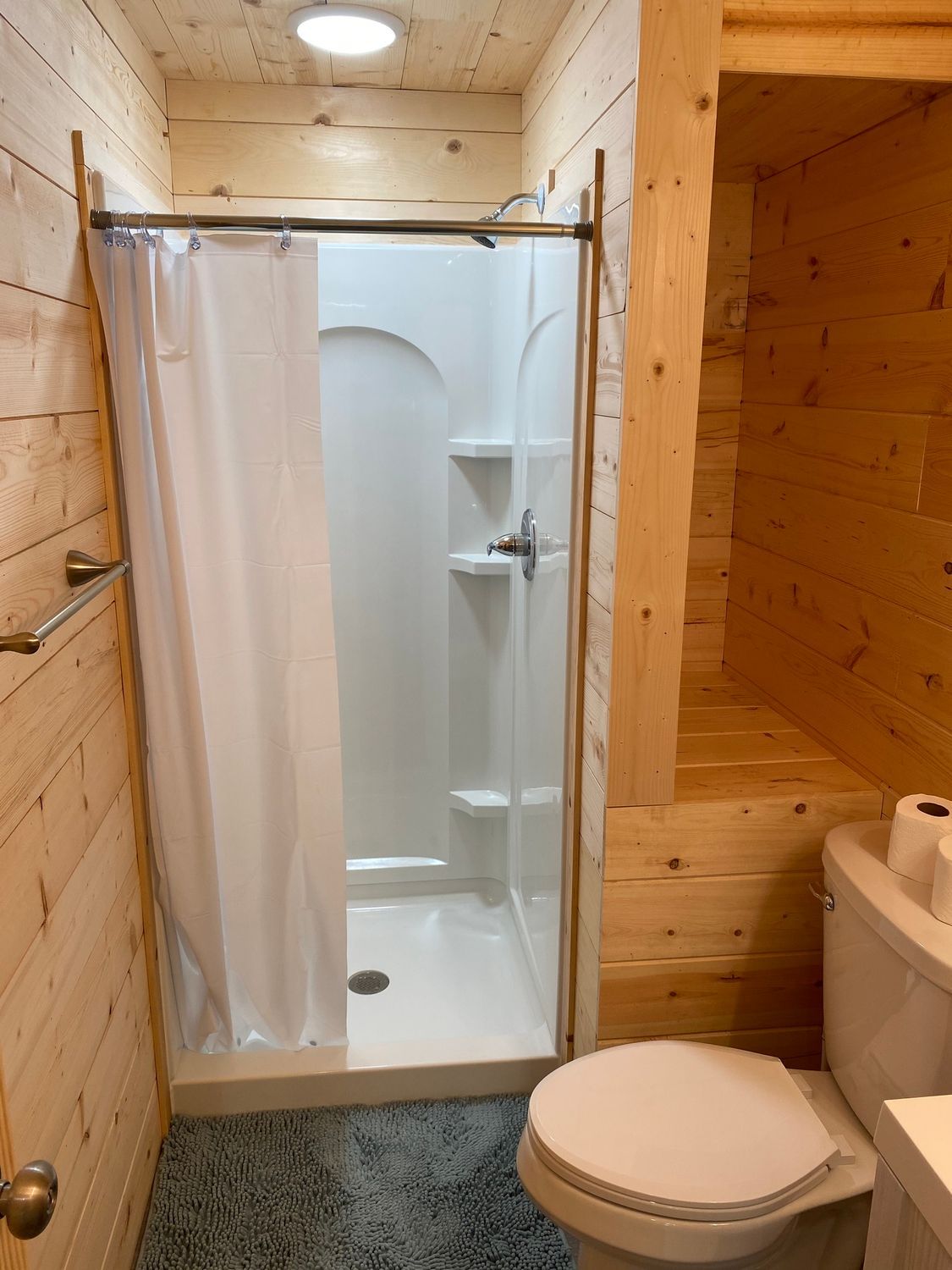 Small wooden-paneled bathroom with a white shower, toilet, and gray speckled flooring.