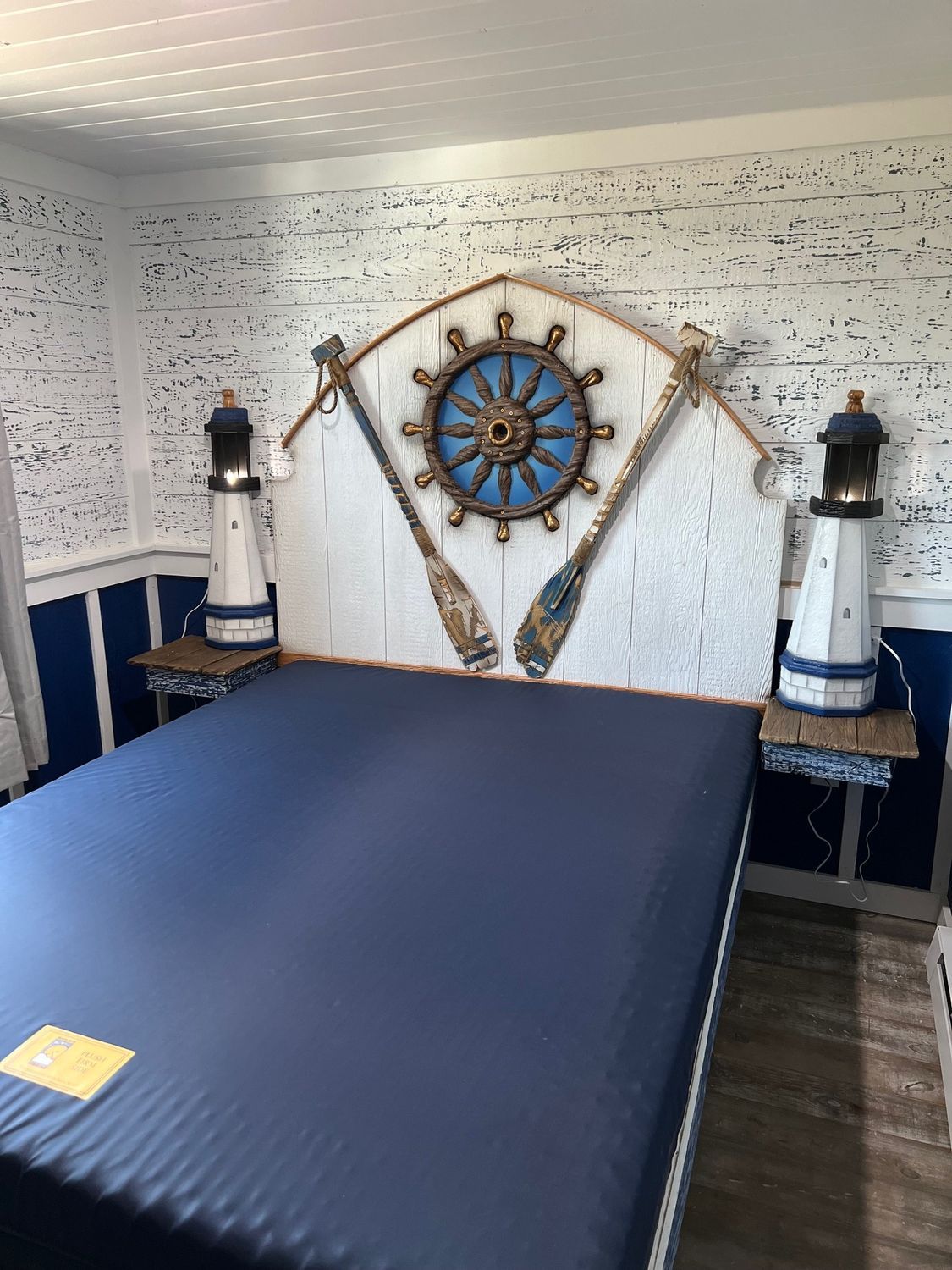 Nautical-themed bedroom with bed, headboard, oars, ship wheel, and lighthouse bedside lamps. Blue and white color scheme.