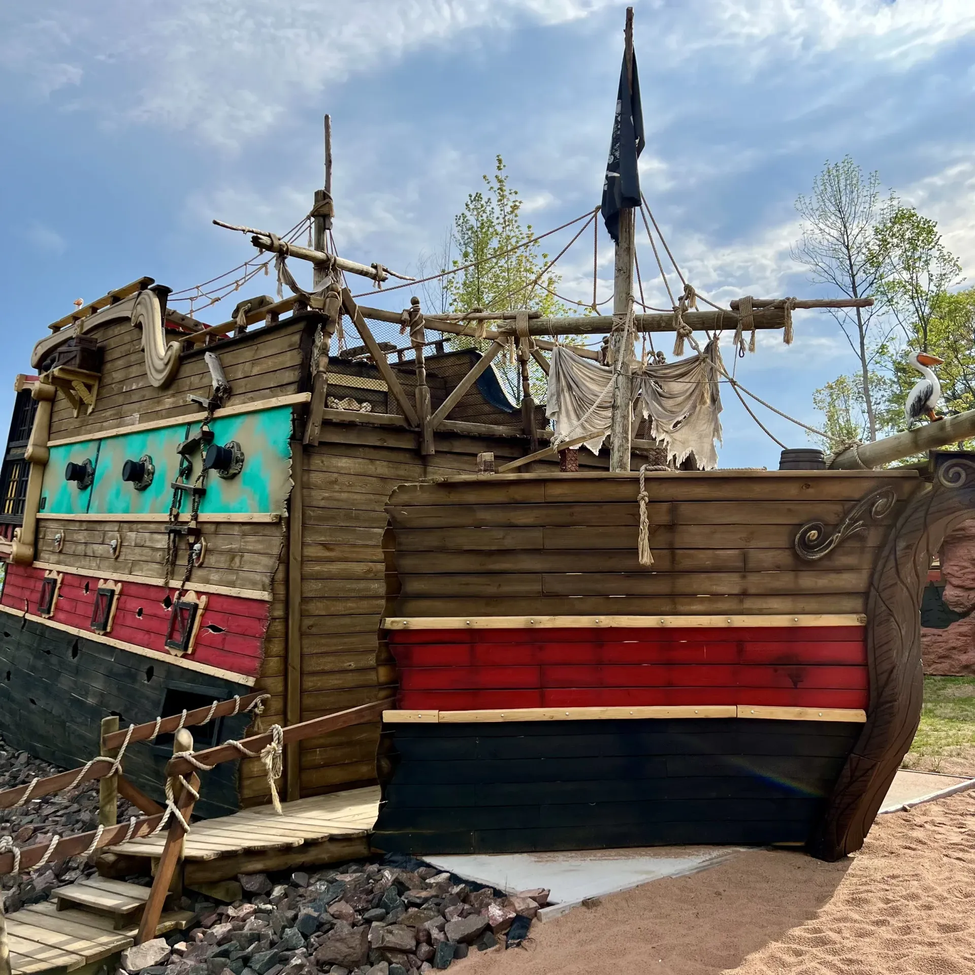 A pirate ship playground structure made of wood, painted with teal, red, and black, set on gravel and sand.