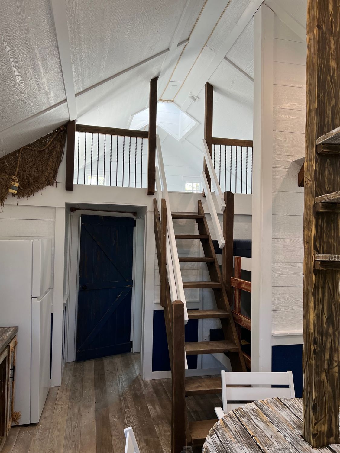 Interior of a cabin with a loft; a staircase leads to the second floor, blue door, white walls, and a white refrigerator.
