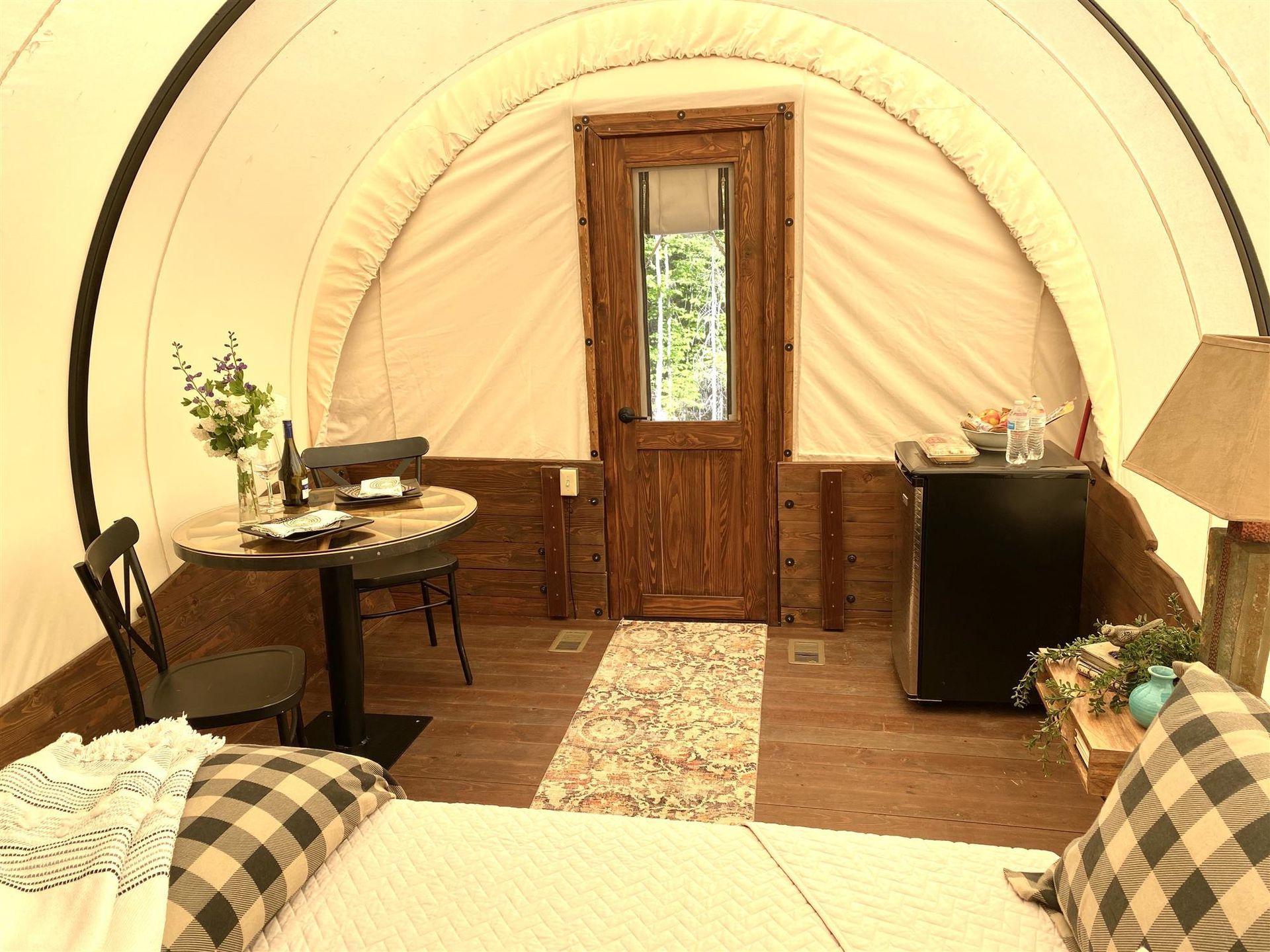 Interior of a glamping wagon. Wooden door and table with flowers, bed, mini-fridge and rug. Beige walls, brown paneling.