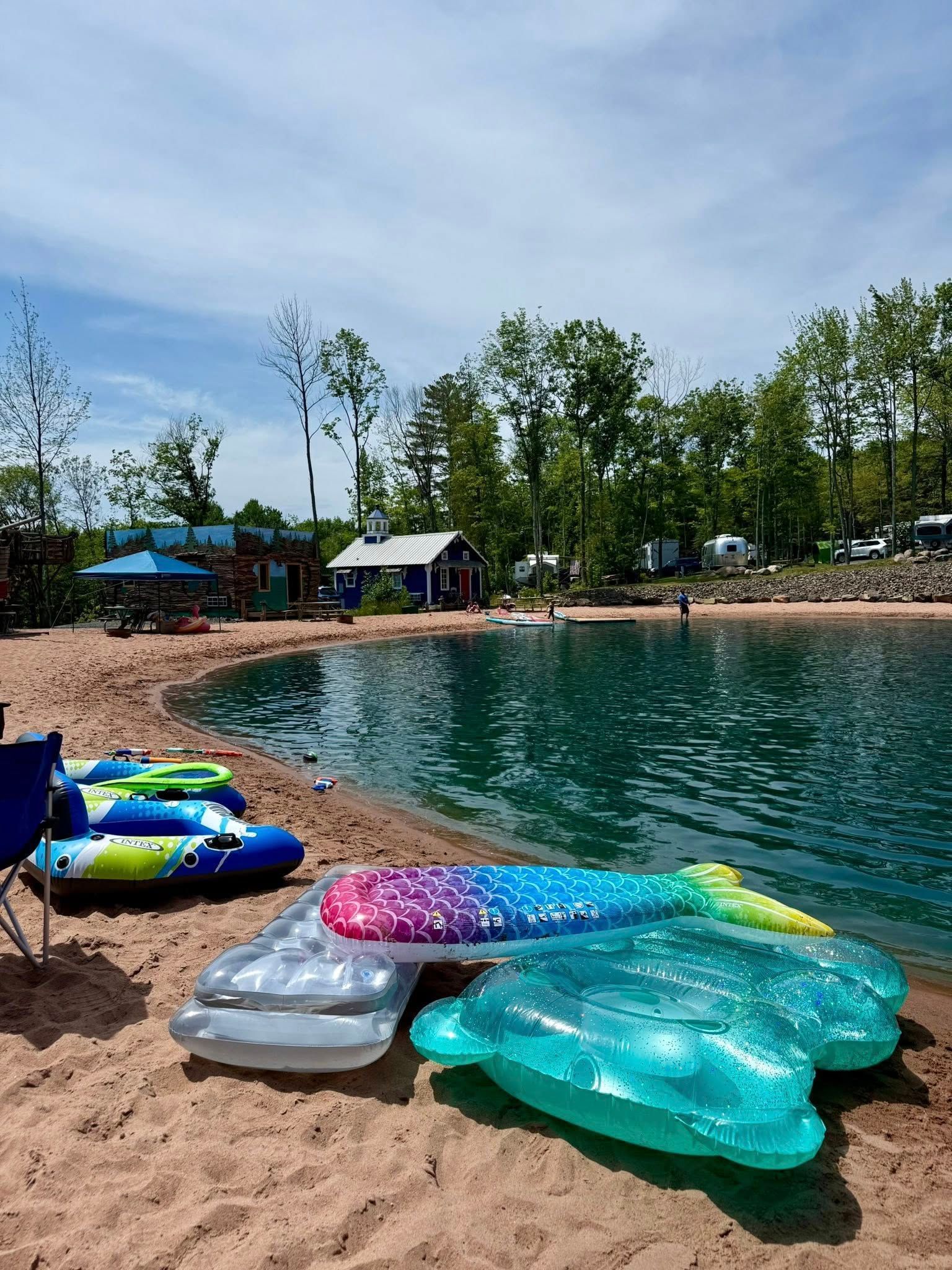 Beach scene: Inflatable floats on sand near blue water. Trees, small buildings, and blue sky in background.