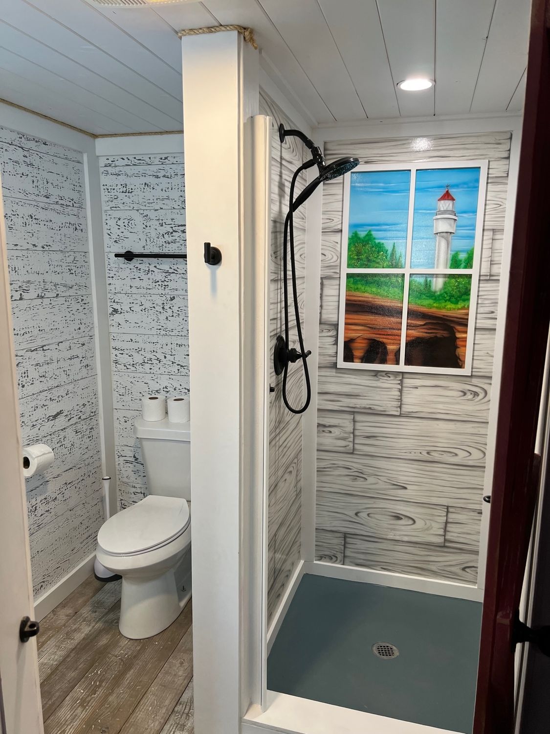 Small bathroom with shower, toilet, and lighthouse painting. White paneling, dark gray fixtures, and blue shower floor.