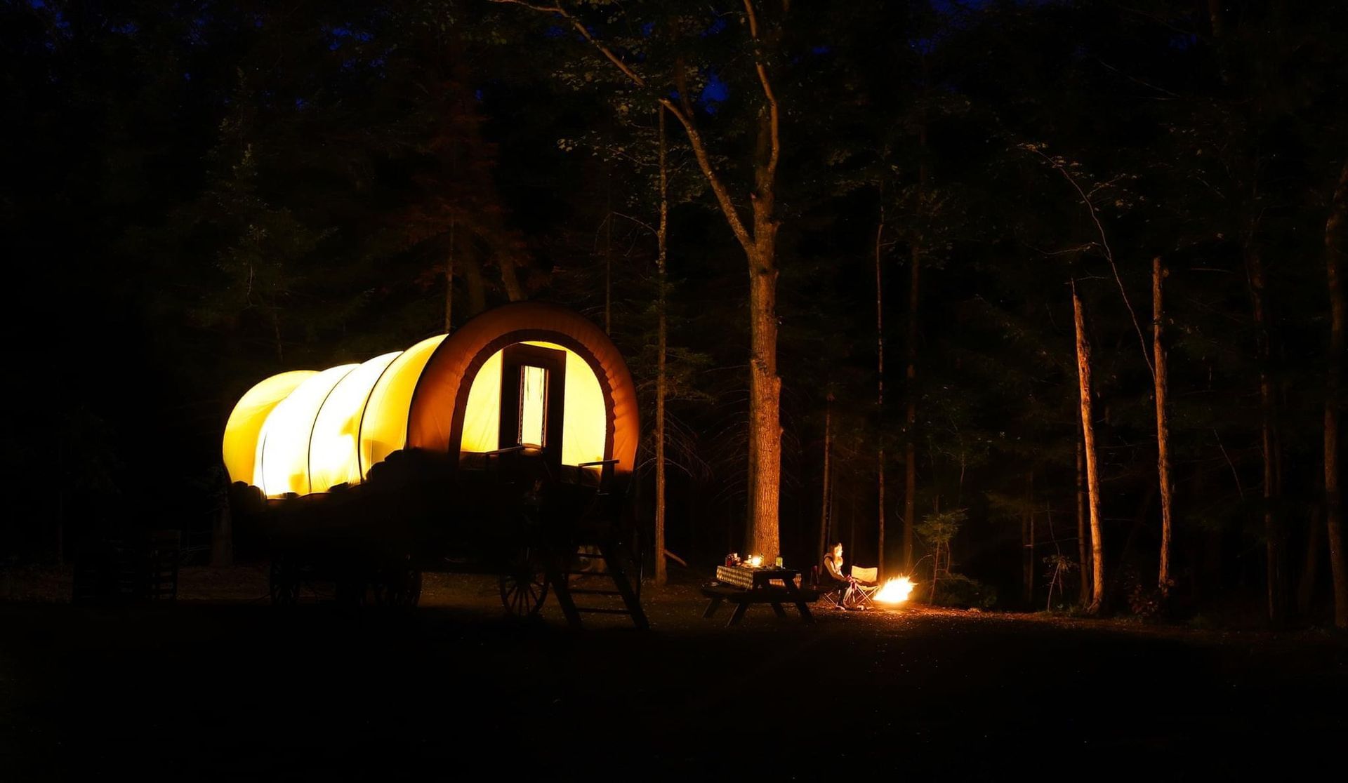 Glowing wagon in a dark forest, with a lit interior and small table set outside.