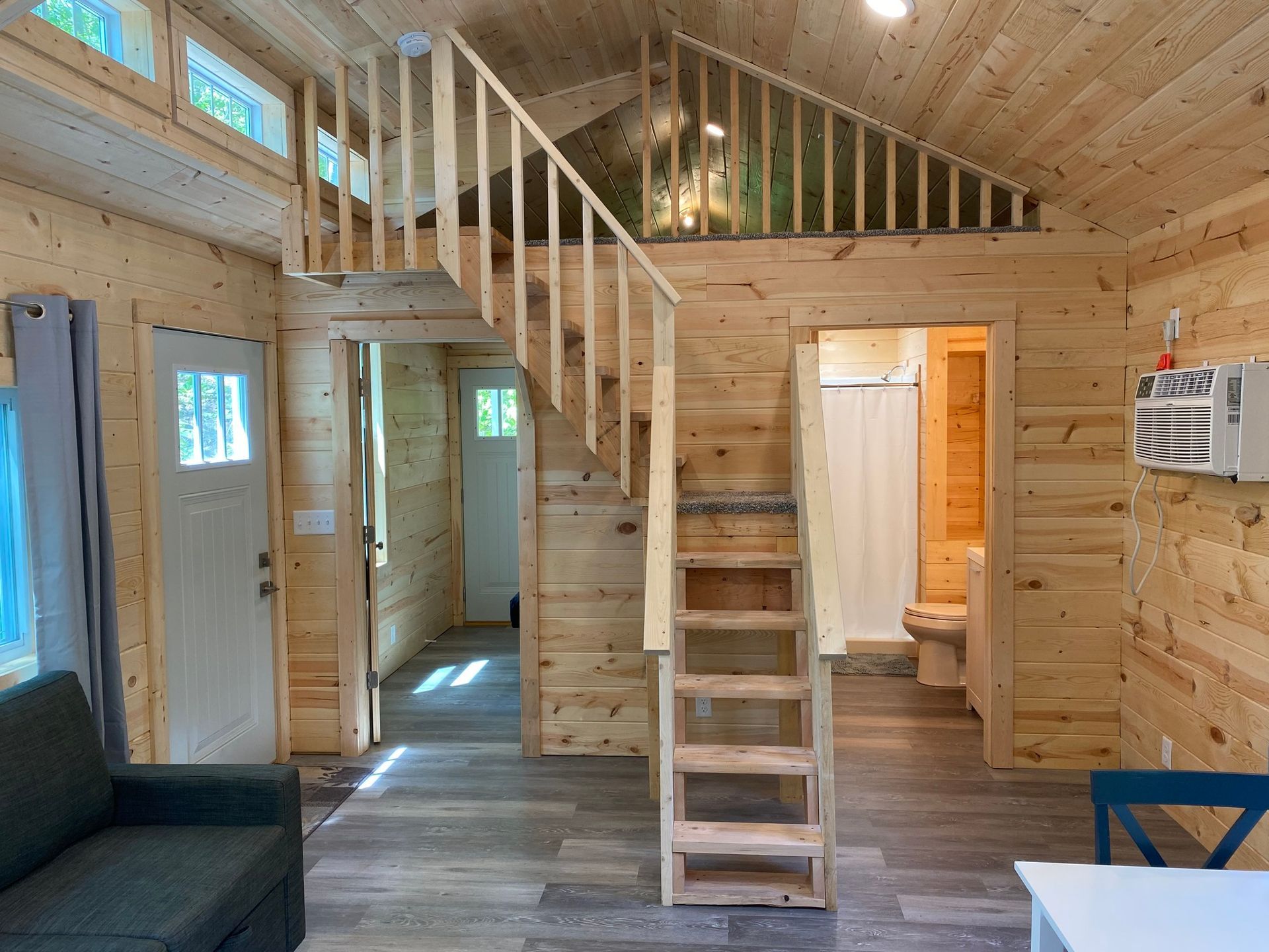 Interior of a tiny cabin: wood walls, stairs to a loft, doorway to bathroom, sofa, window.