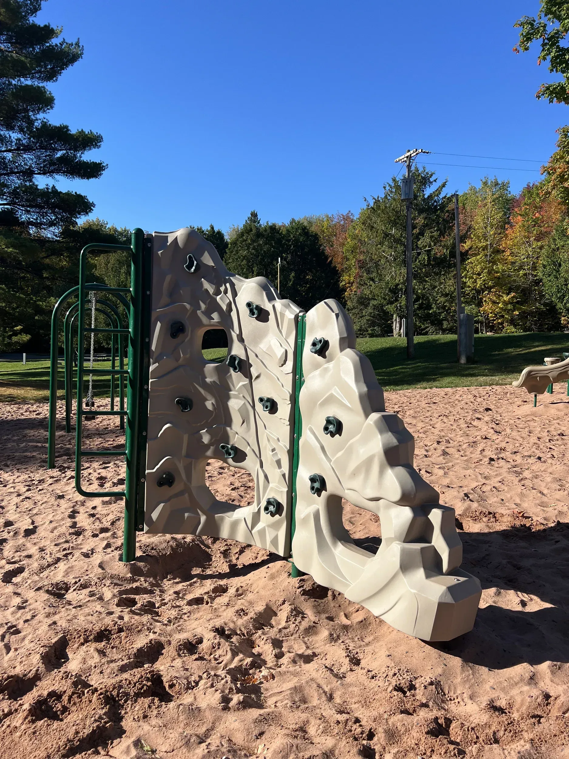 Climbing wall structure in a sandy playground, green metal frame, tan climbing surface with black holds, clear blue sky.