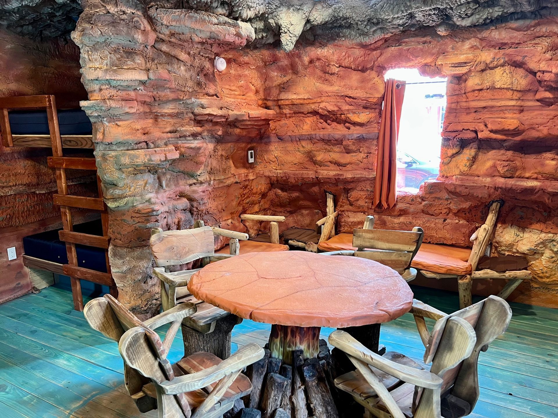 Cave-like room with round table and rustic chairs. Bunk beds on left, window with curtains on right.