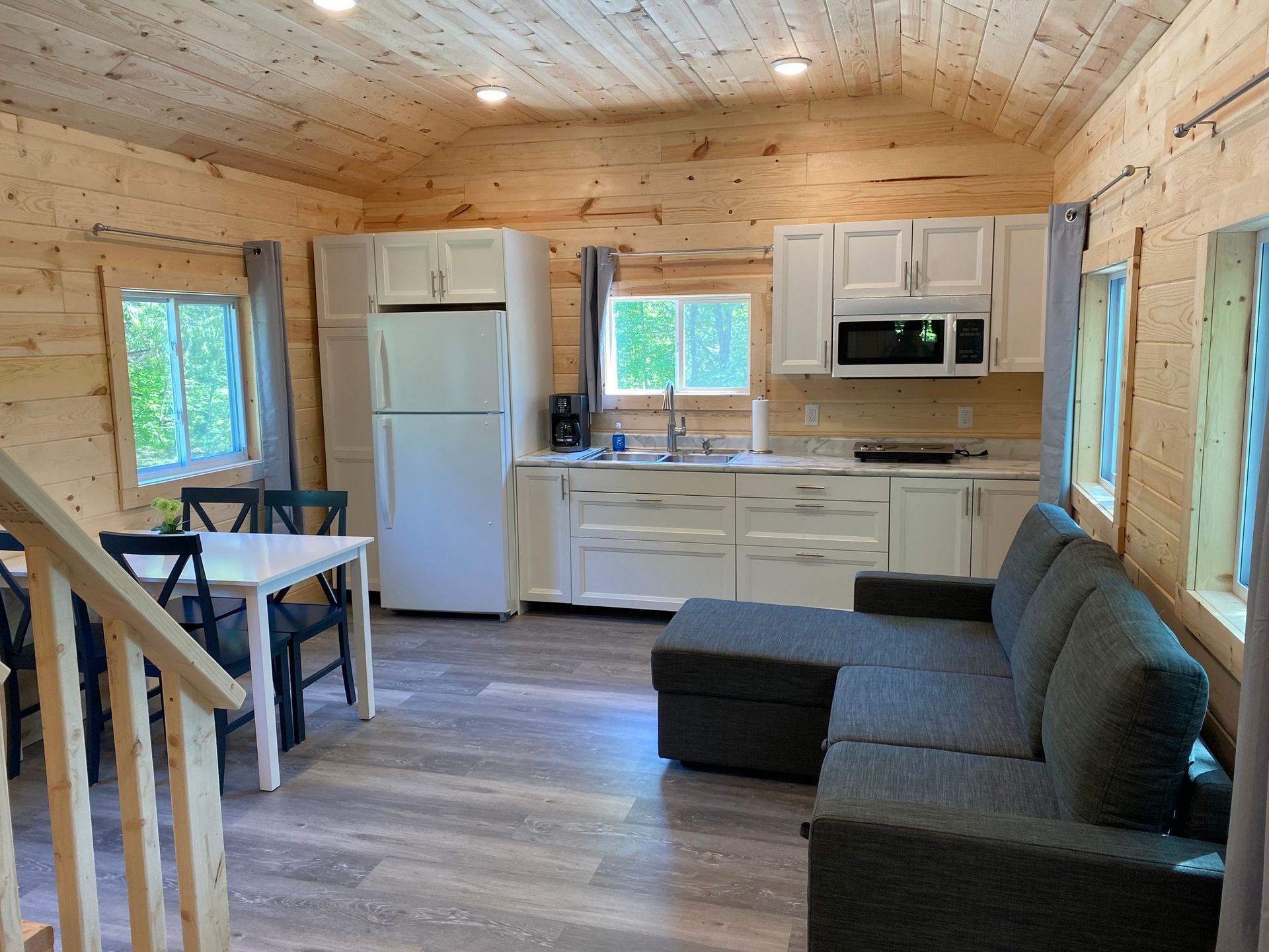 Interior cabin with kitchen, dining area, and gray sectional sofa. White cabinets and fridge. Wooden walls.