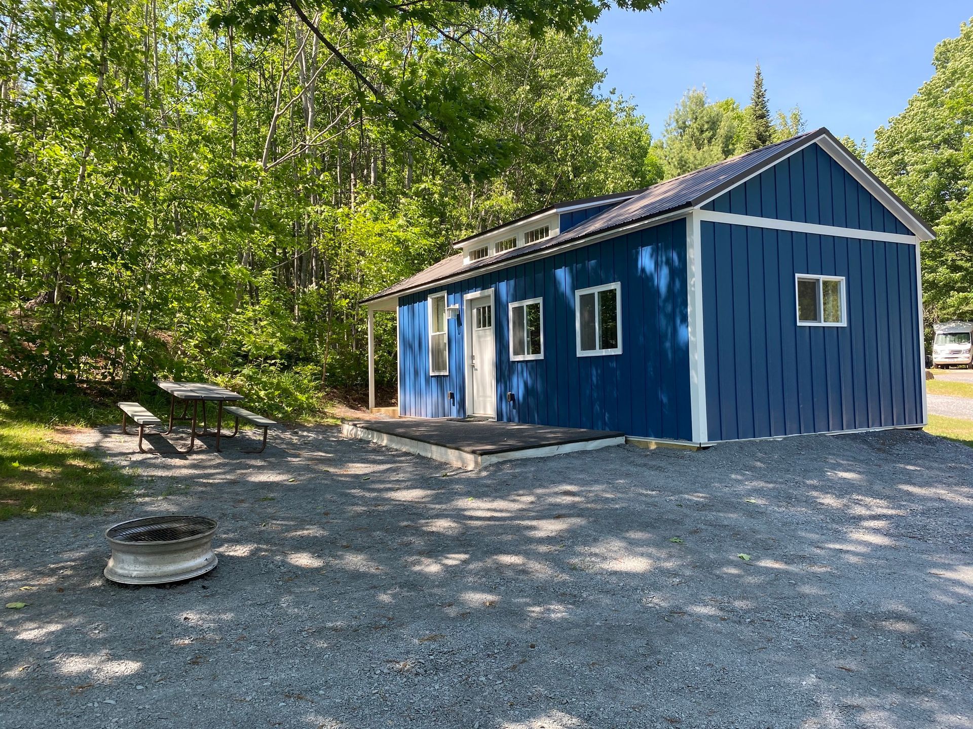Blue cabin with white trim, picnic table, and fire pit in a gravel area surrounded by trees.