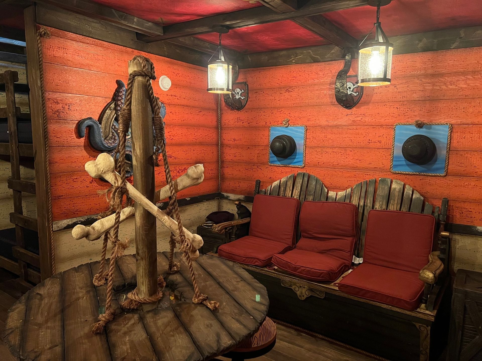 Cozy pirate-themed room with red walls, bench seating, hanging lanterns, and a wooden table with hanging bones.