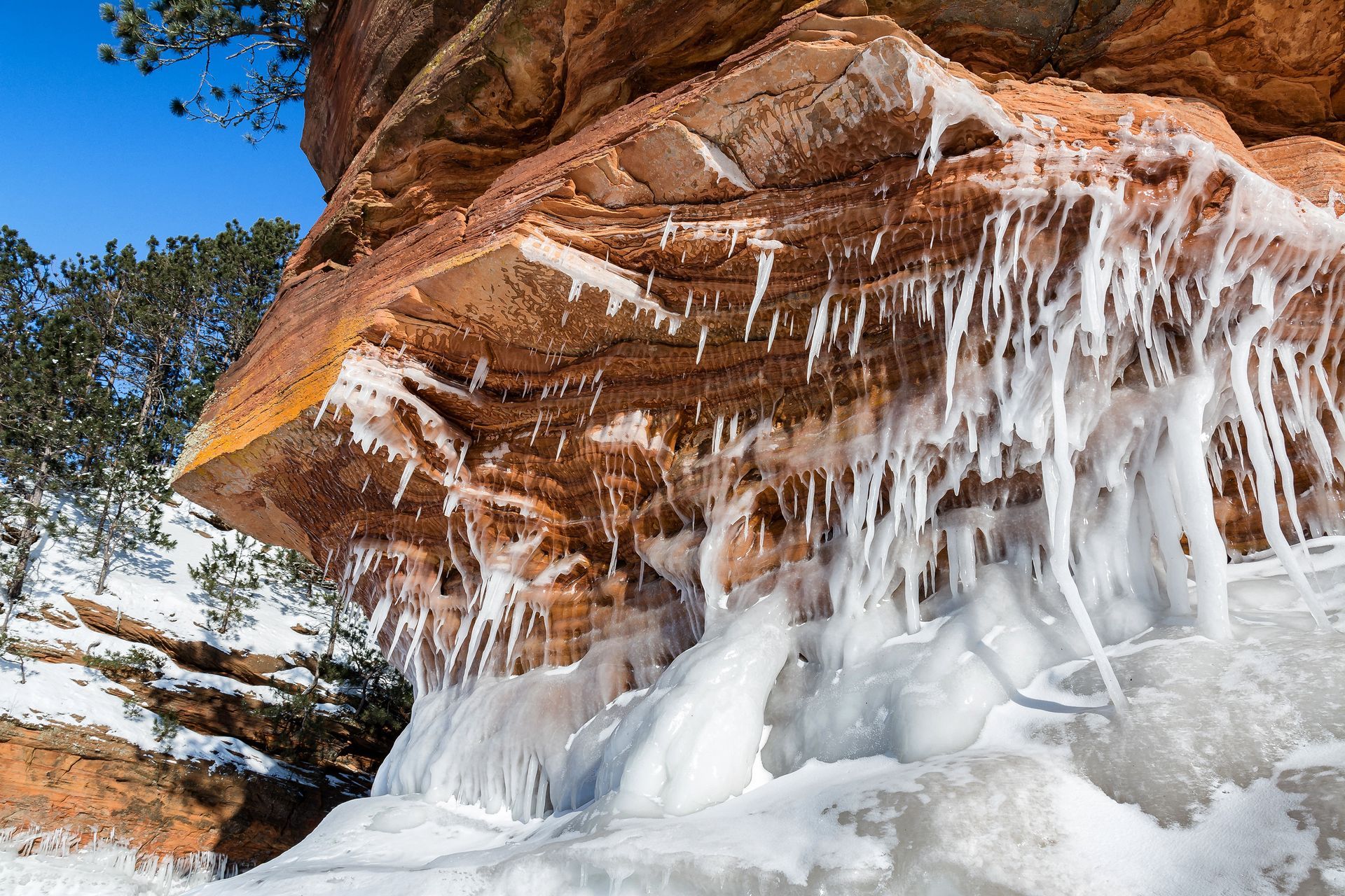 Ice formations and icicles hanging from a weathered orange sandstone cliff face against a clear blue sky.