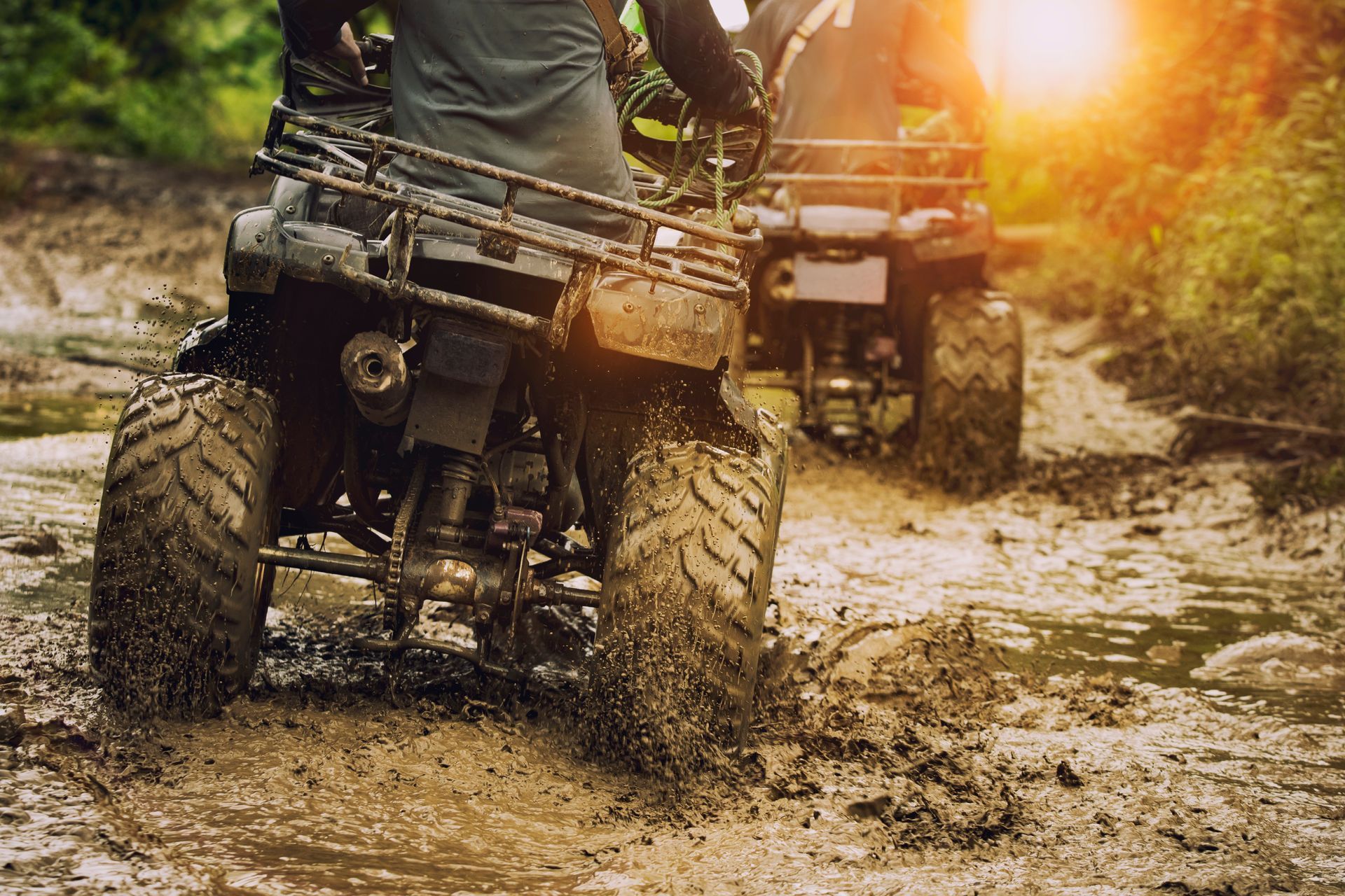 ATVs driving through muddy terrain. Sun shining, tires splashing mud.