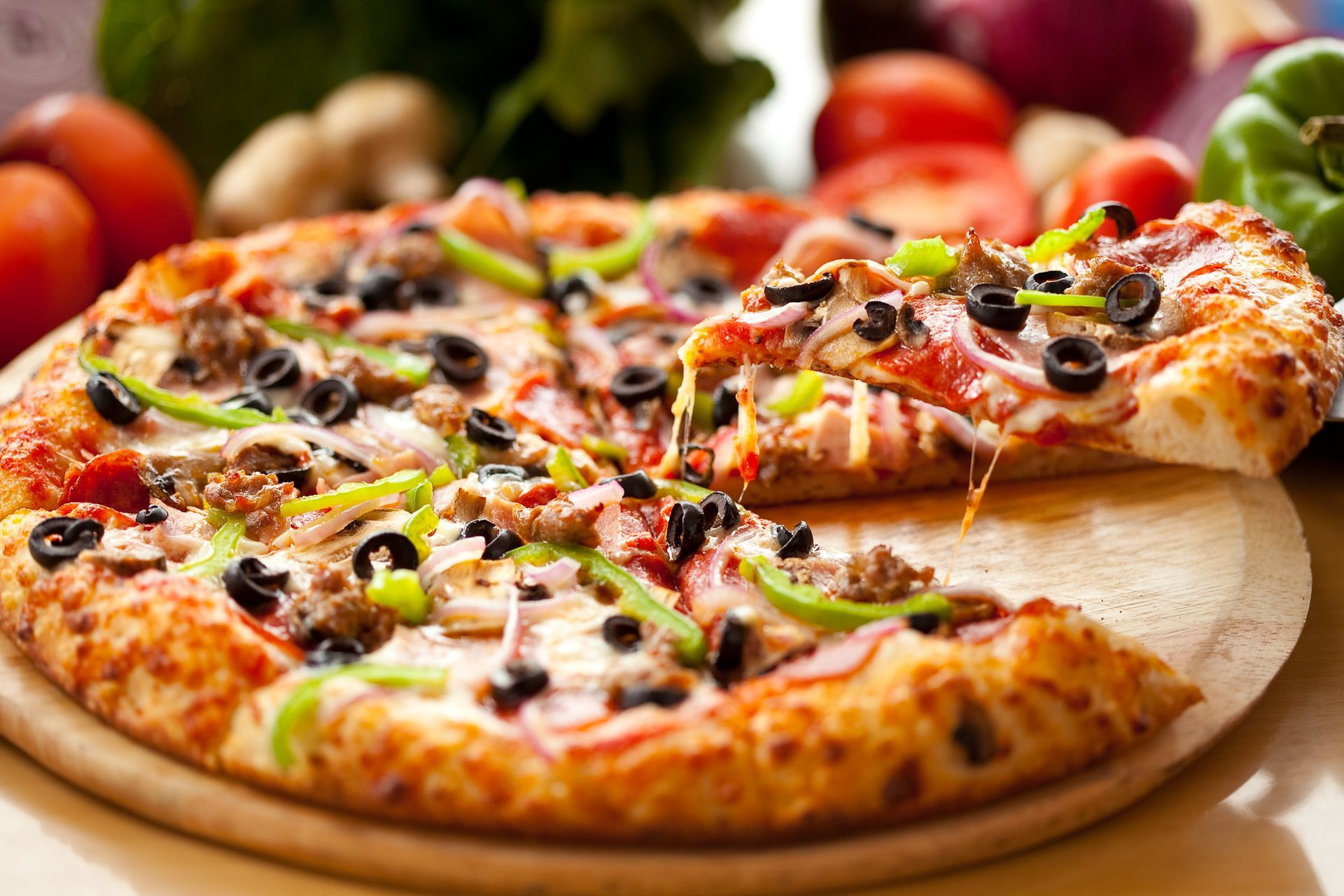 Pizza with melted cheese, black olives, and green peppers on a wooden board. A slice is being lifted.