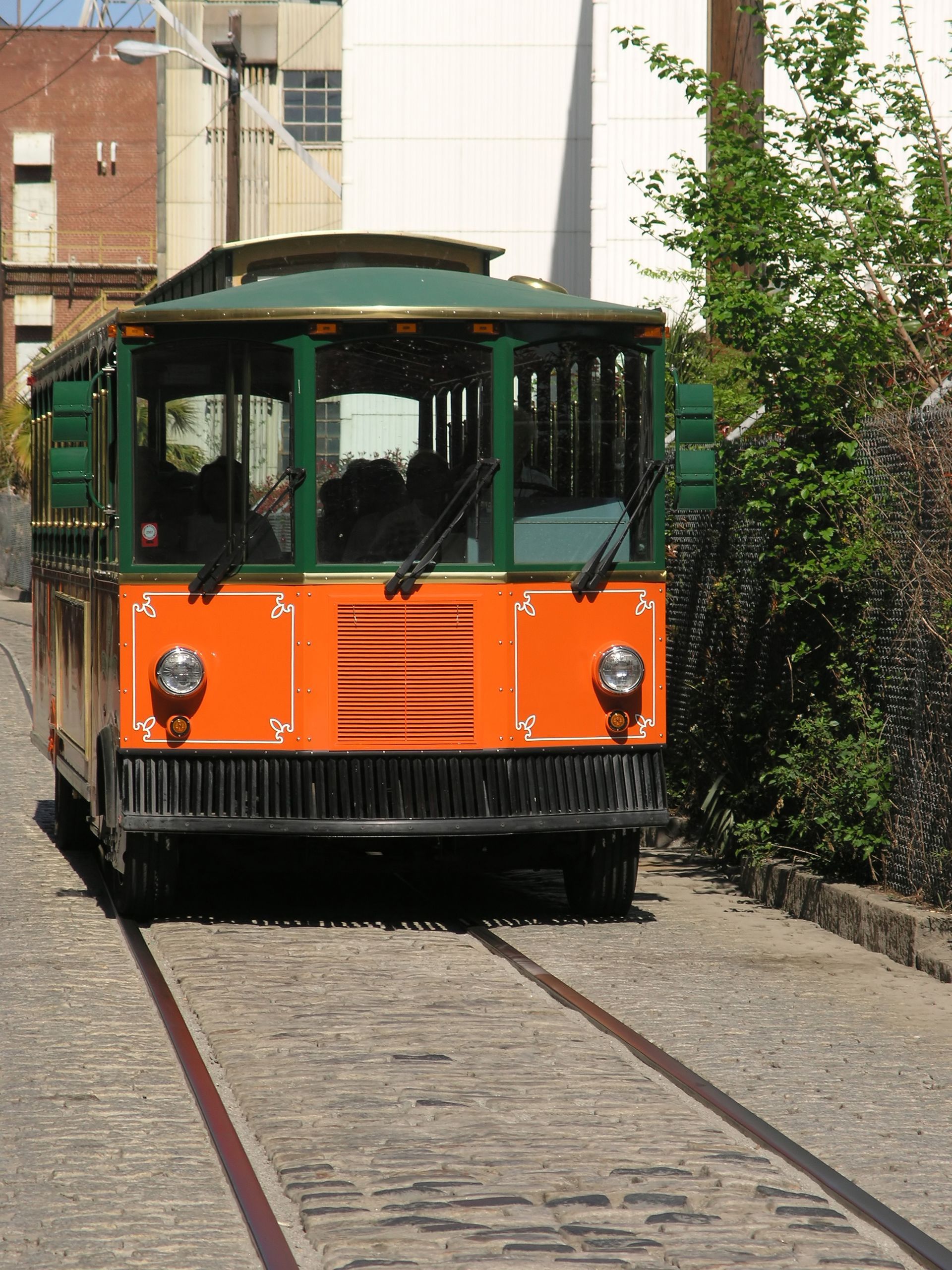 Orange and green trolley on tracks, brick street, buildings in background.