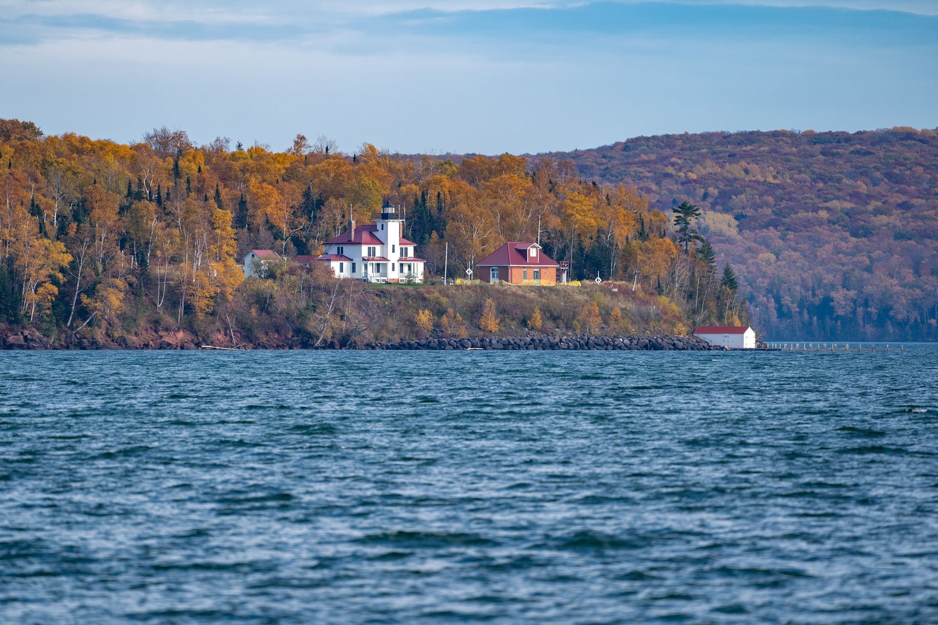 Lakeside cliff with autumn trees, houses, and a sailboat on calm blue water