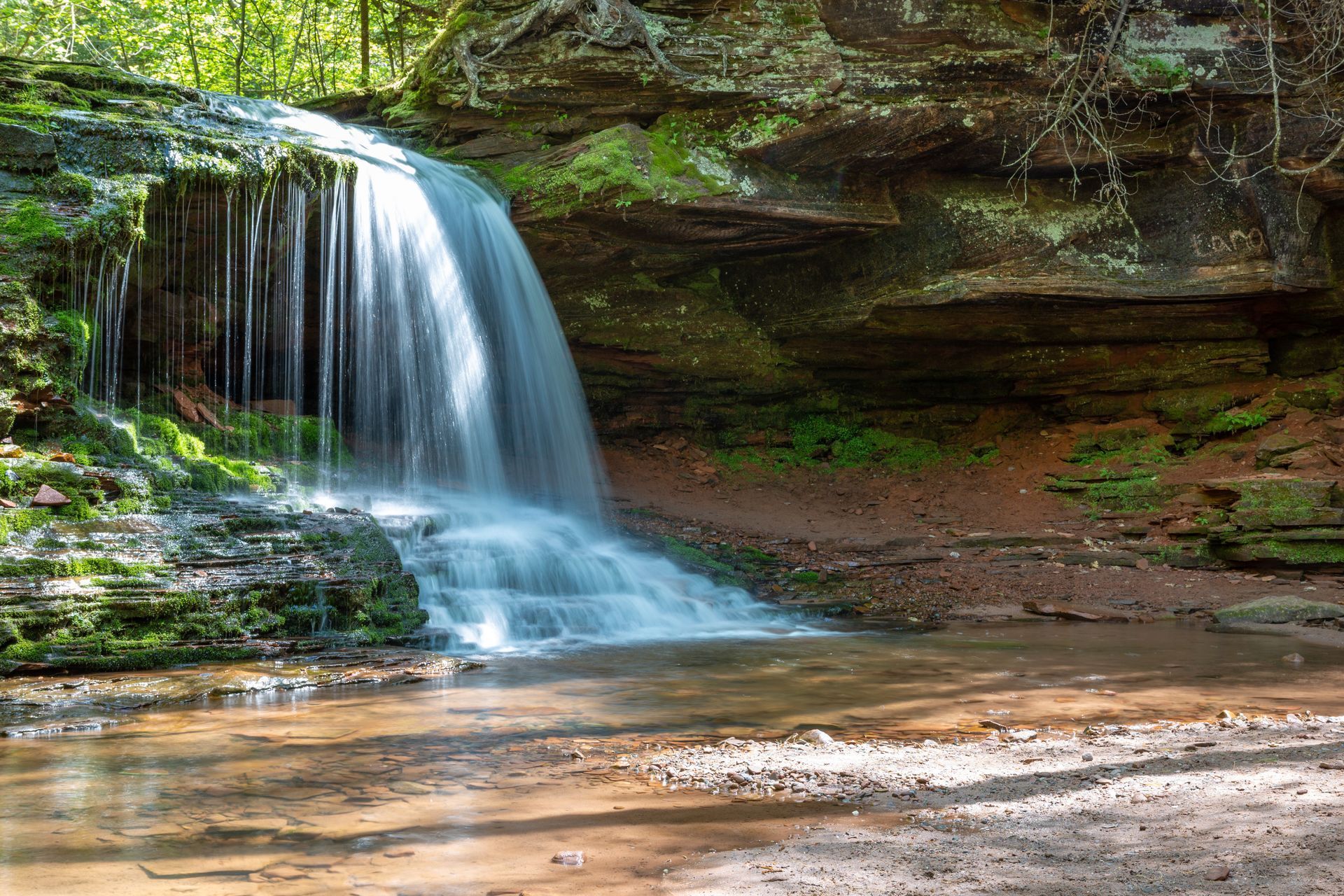 Waterfall cascading into a clear pool, surrounded by mossy rocks and a forest setting.