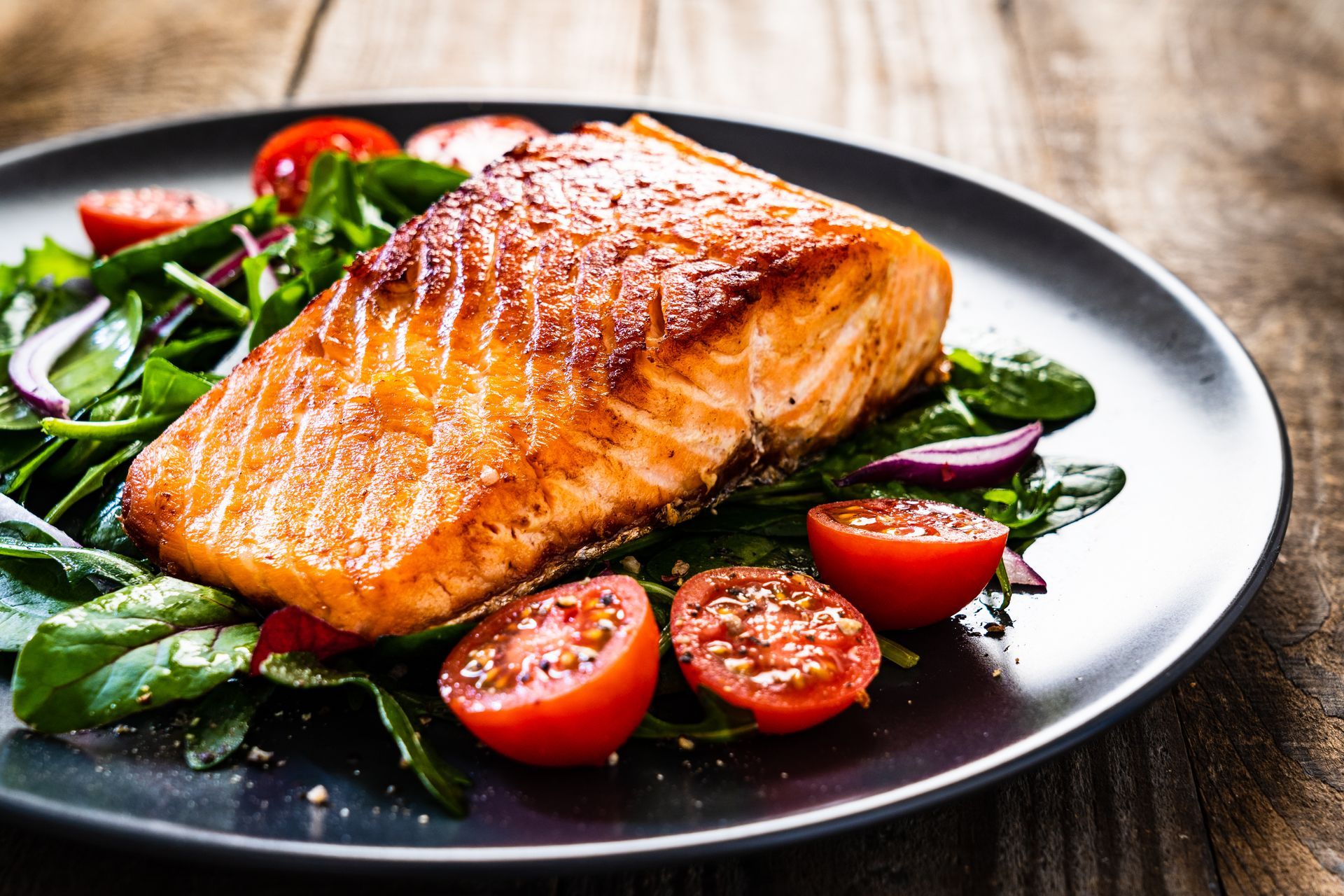 Seared salmon fillet on a bed of spinach, tomatoes, and red onion, on a dark plate.