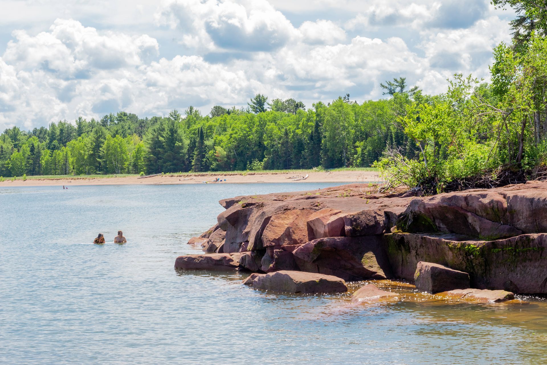 Two people swim in a blue lake next to a red sandstone rocky outcrop, with a sandy beach and lush forest in the back.