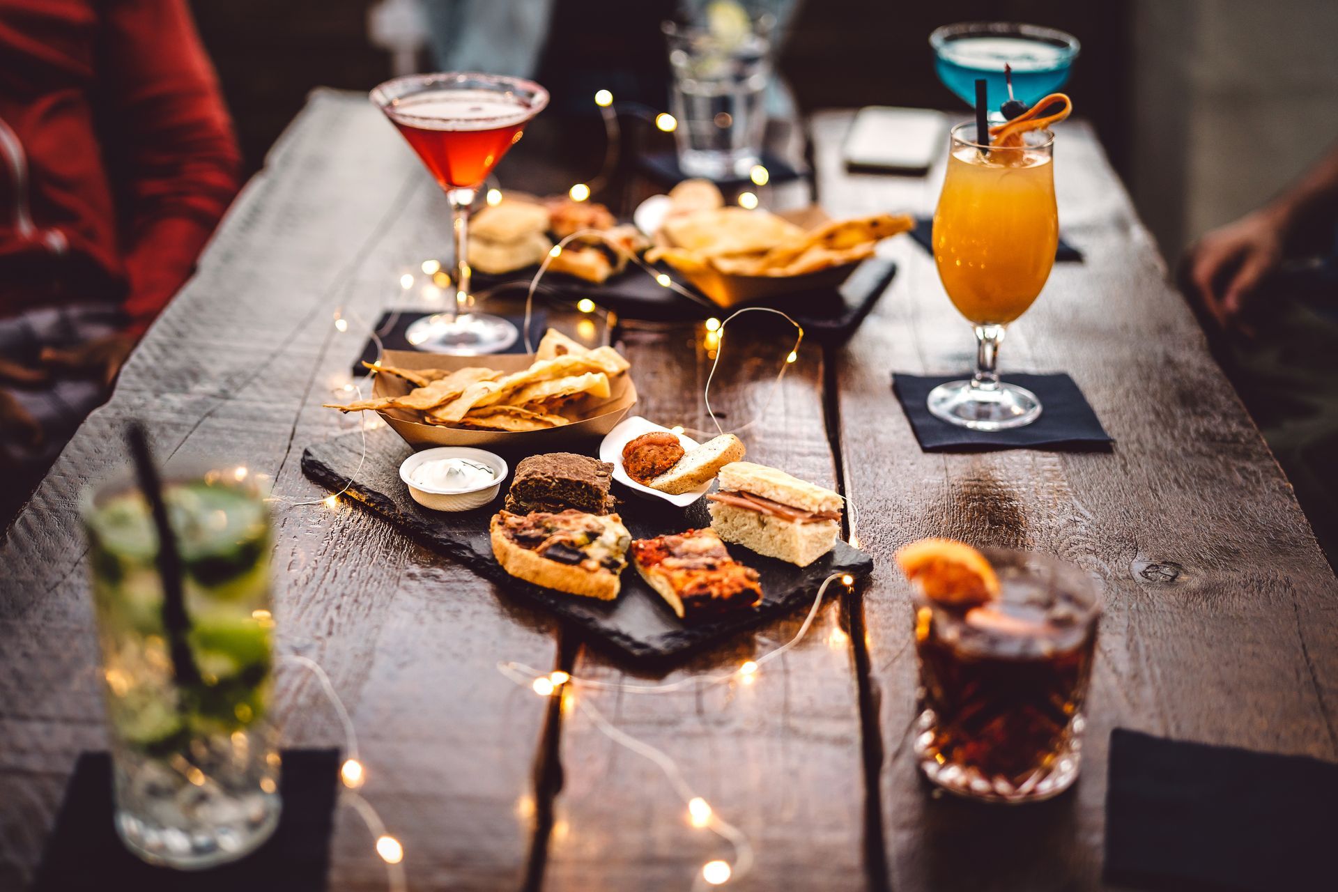 An assortment of drinks and snacks on a dark wooden table lit by string lights.
