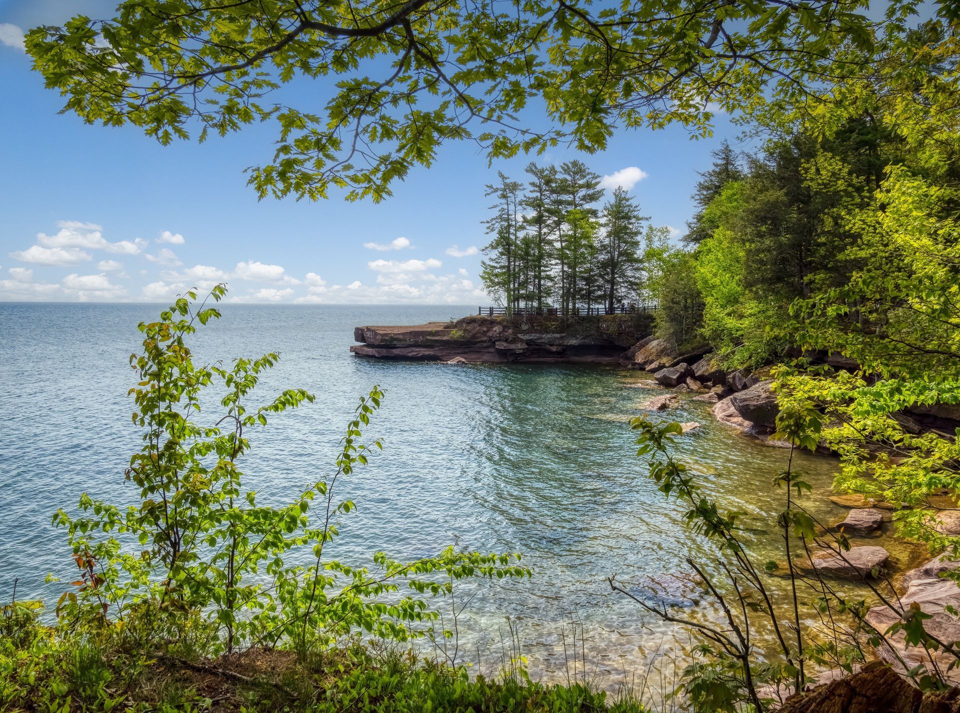Lush green trees frame a blue lake under a partly cloudy sky, rocky shoreline with trees in the distance.