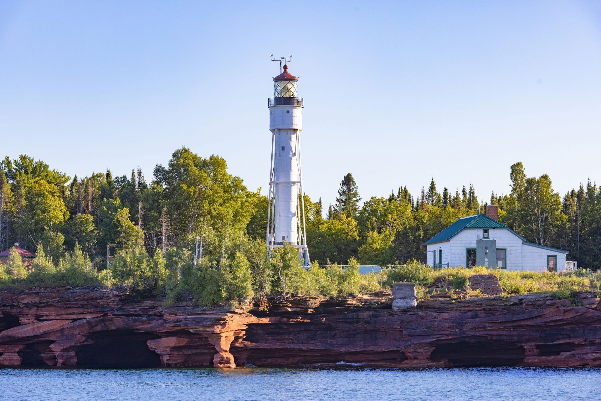 A tall, white lighthouse stands on a red rocky cliff next to a white building, set against a backdrop of green trees.