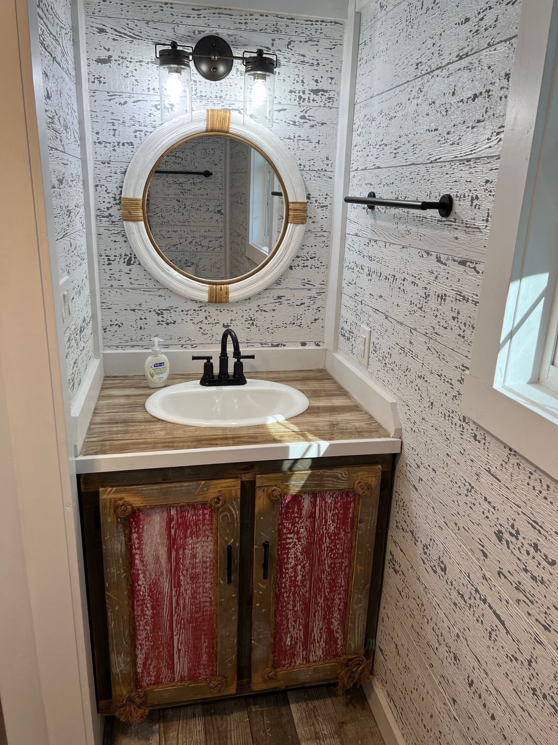 Rustic bathroom with white shiplap walls, wood vanity, and round mirror with gold trim.