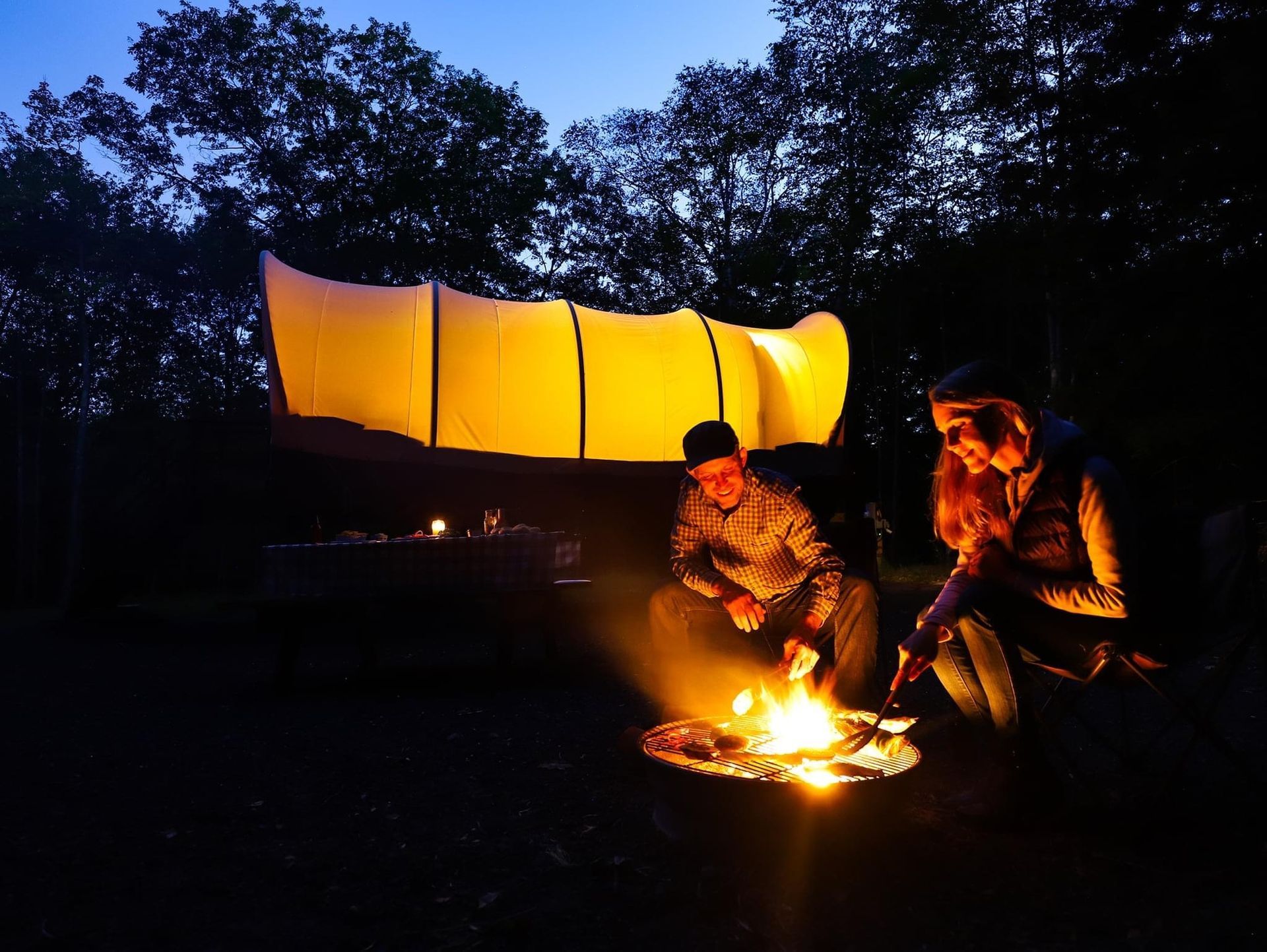 Two people by a campfire at dusk, firelight illuminating faces. A glowing white structure is behind them.