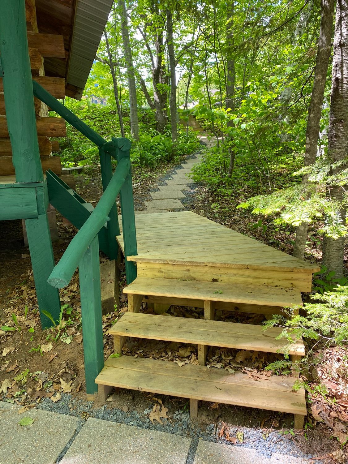 Wooden stairs leading to a path in a wooded area, next to a green-painted log structure.