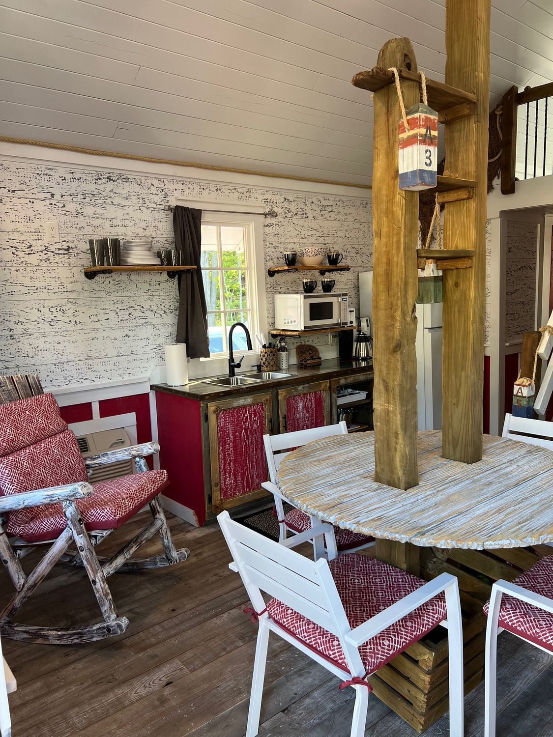 Interior view of a rustic kitchen with a round table and chairs. Red and white accents. Wooden post with shelves.
