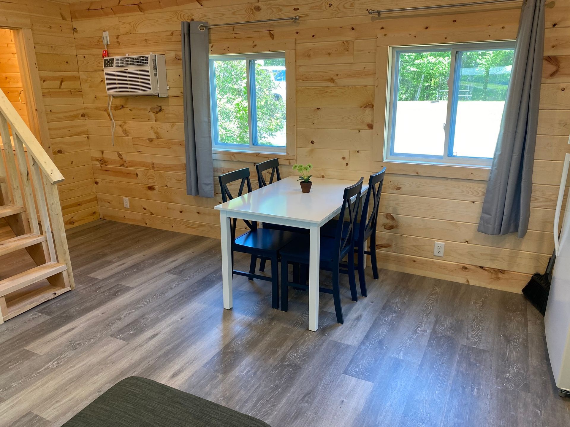 Interior of a cabin with a dining table, chairs, windows, and wooden walls. Grey flooring, white table.