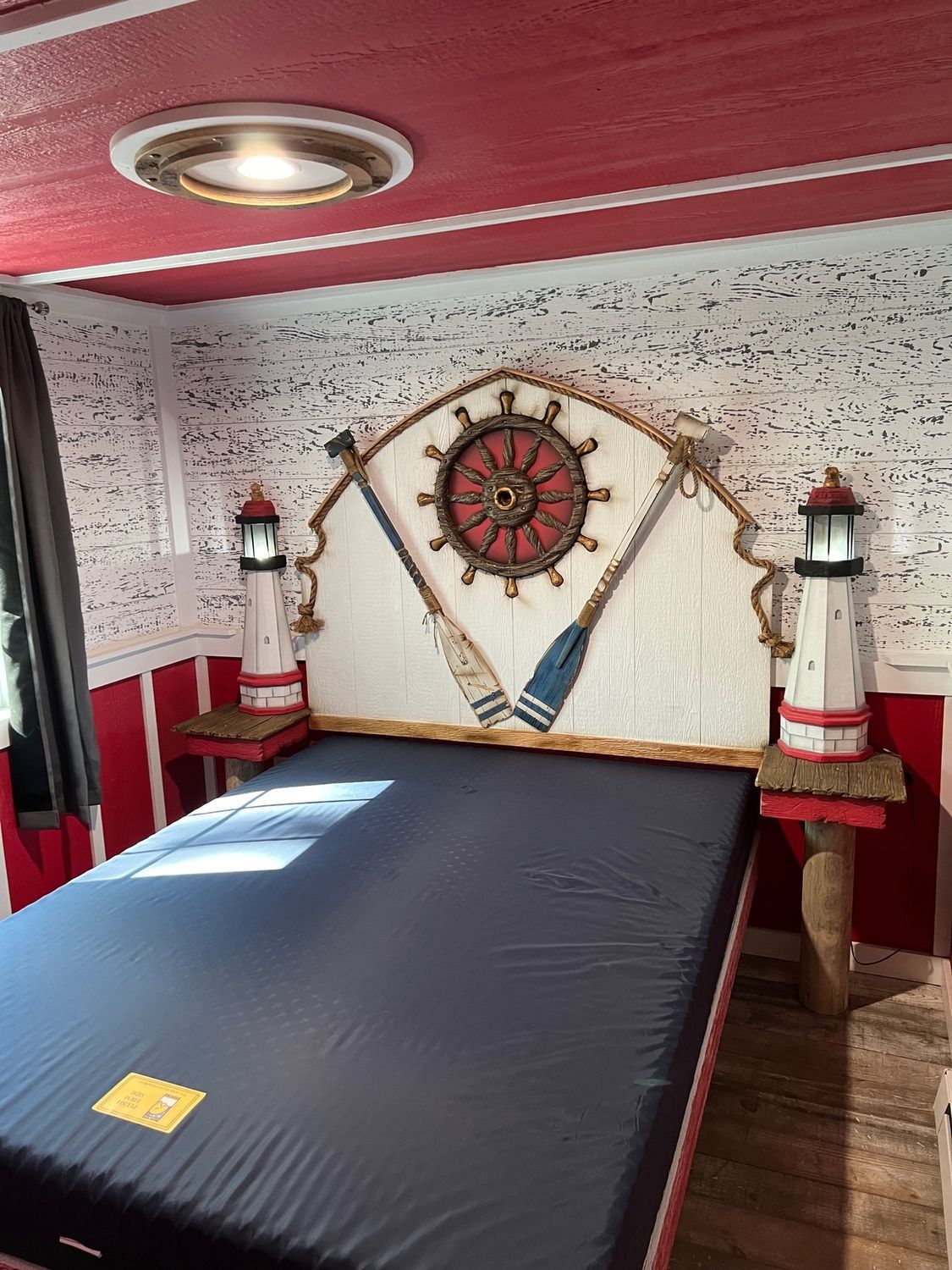 Nautical-themed bedroom with a bed, a ship wheel headboard, lighthouse lamps, and red and white walls.