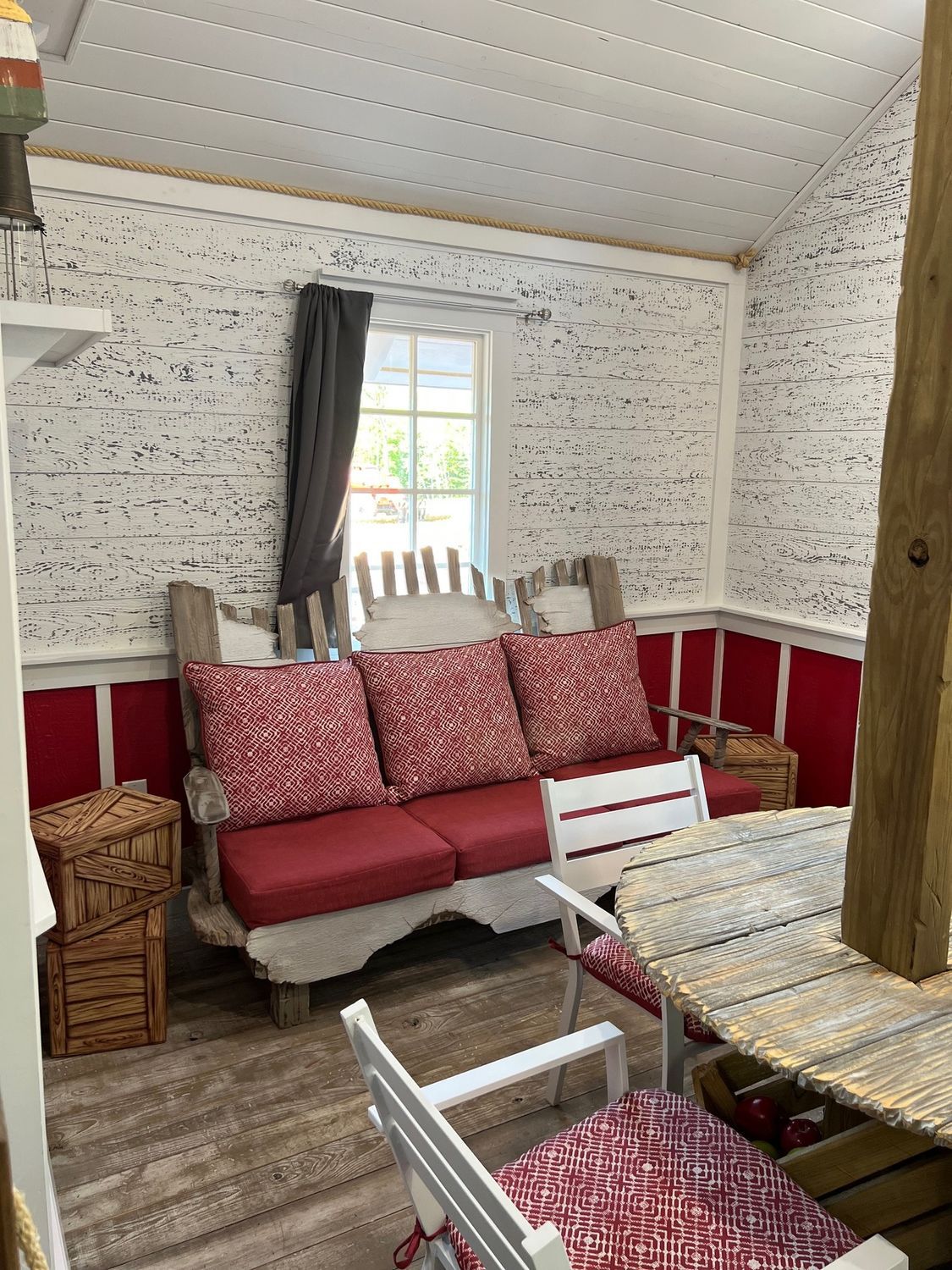 Cozy enclosed outdoor space with red and white decor: couch, chairs, window, wooden crate and table.