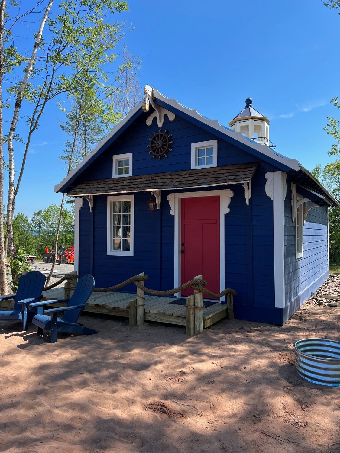 Blue cabin with red door and small lighthouse top, wood trim, two Adirondack chairs outside.