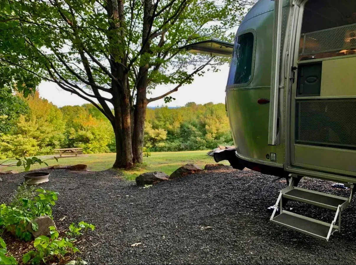 Silver camper trailer parked near a tree, gravel ground, fall foliage in background.