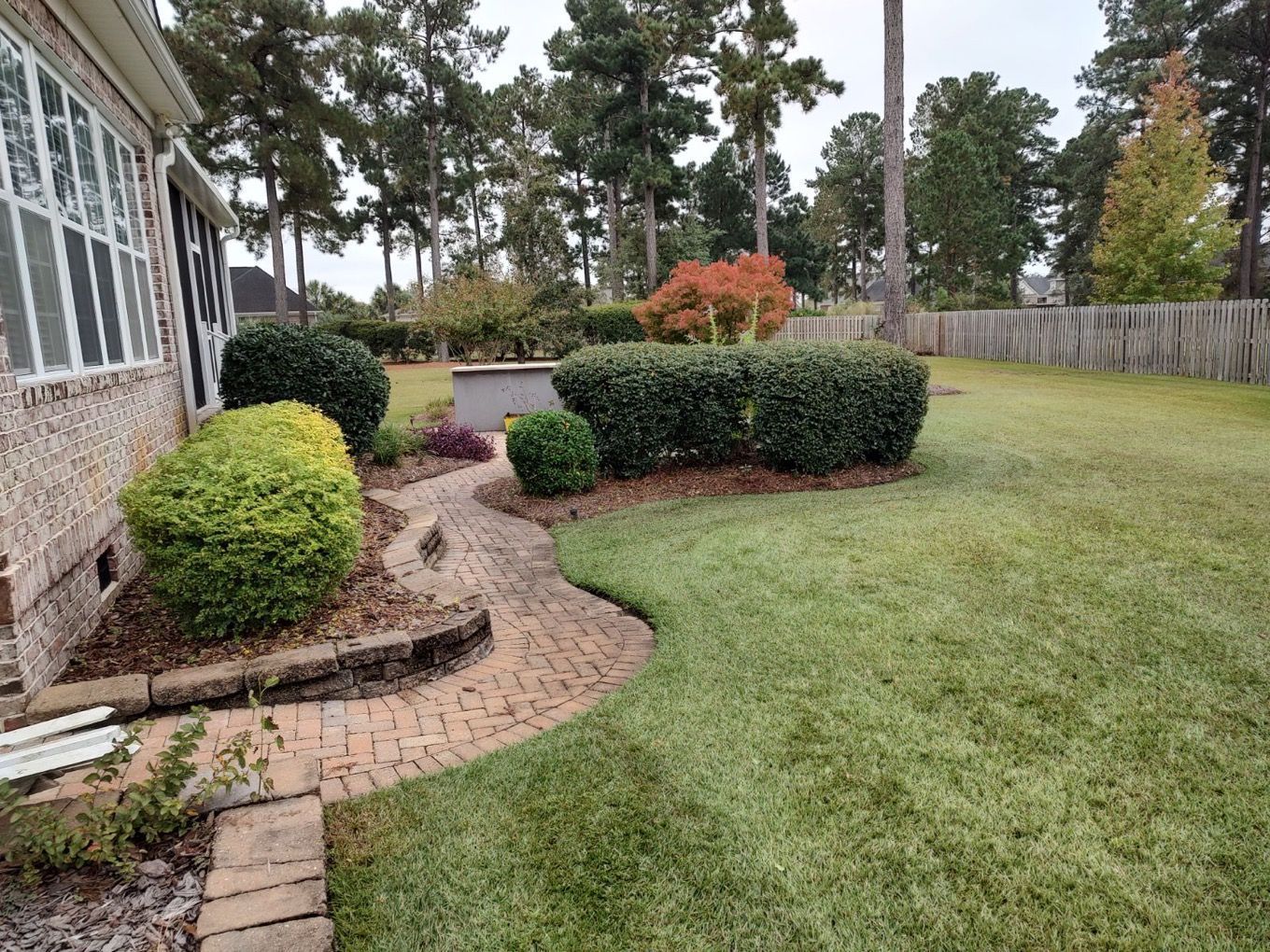 Curved brick walkway beside a house with manicured shrubs and a green lawn under trees