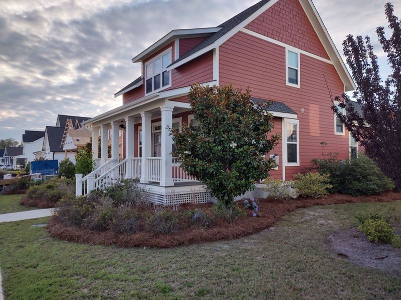 Red two-story house with white trim and porch, surrounded by shrubs and lawn under a cloudy sky