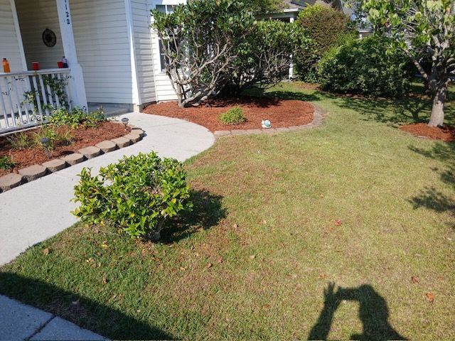 Front yard with curved walkway, trimmed lawn, shrubs, and a house porch in sunlight.