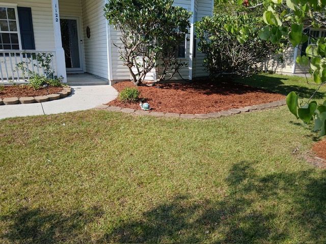 Front yard with lawn, curved mulch beds, shrubs, and a house entryway under bright sunlight.