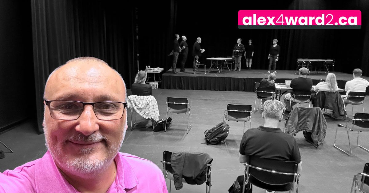 Man smiling in pink shirt at indoor theatre stage with seated audience and alex4ward2.ca sign