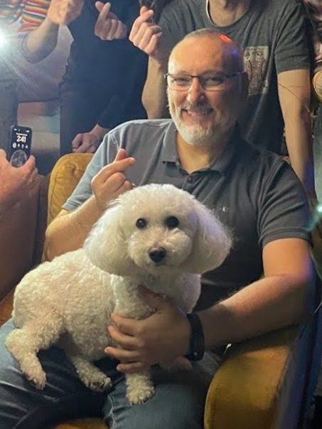 Man sitting in a chair holding a fluffy white dog, smiling at the camera in a crowded indoor setting