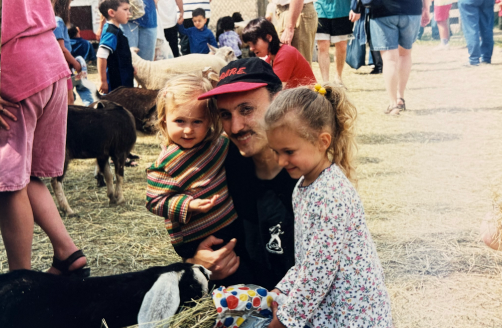 Person in red cap crouches with two children at an outdoor fair, smiling among a crowd.