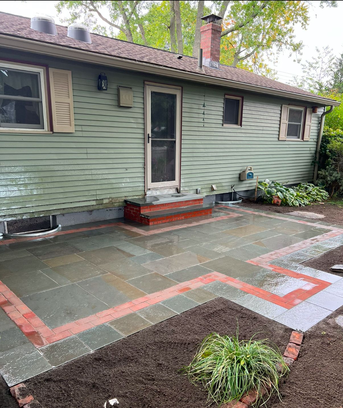 Back patio with green house siding, red-brick steps, and a paved accessible path beside a garden bed