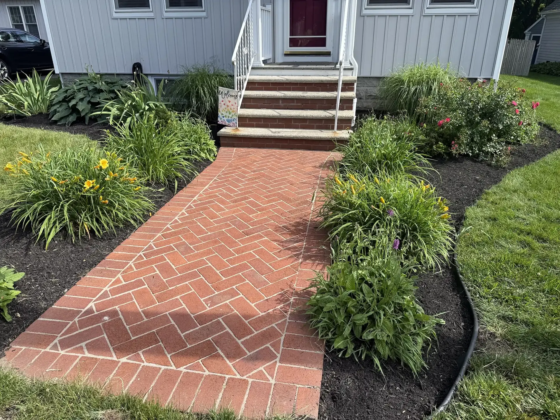 A brick walkway with a herringbone pattern leads to a set of stairs at the entrance of a house, flanked by garden beds.