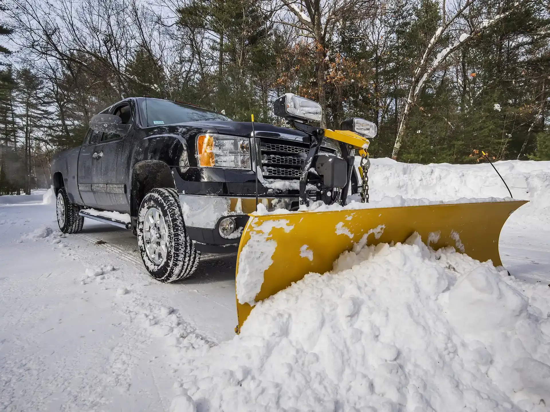 A dark pickup truck with a large yellow snow plow attached to the front, clearing snow in a wooded, winter setting.