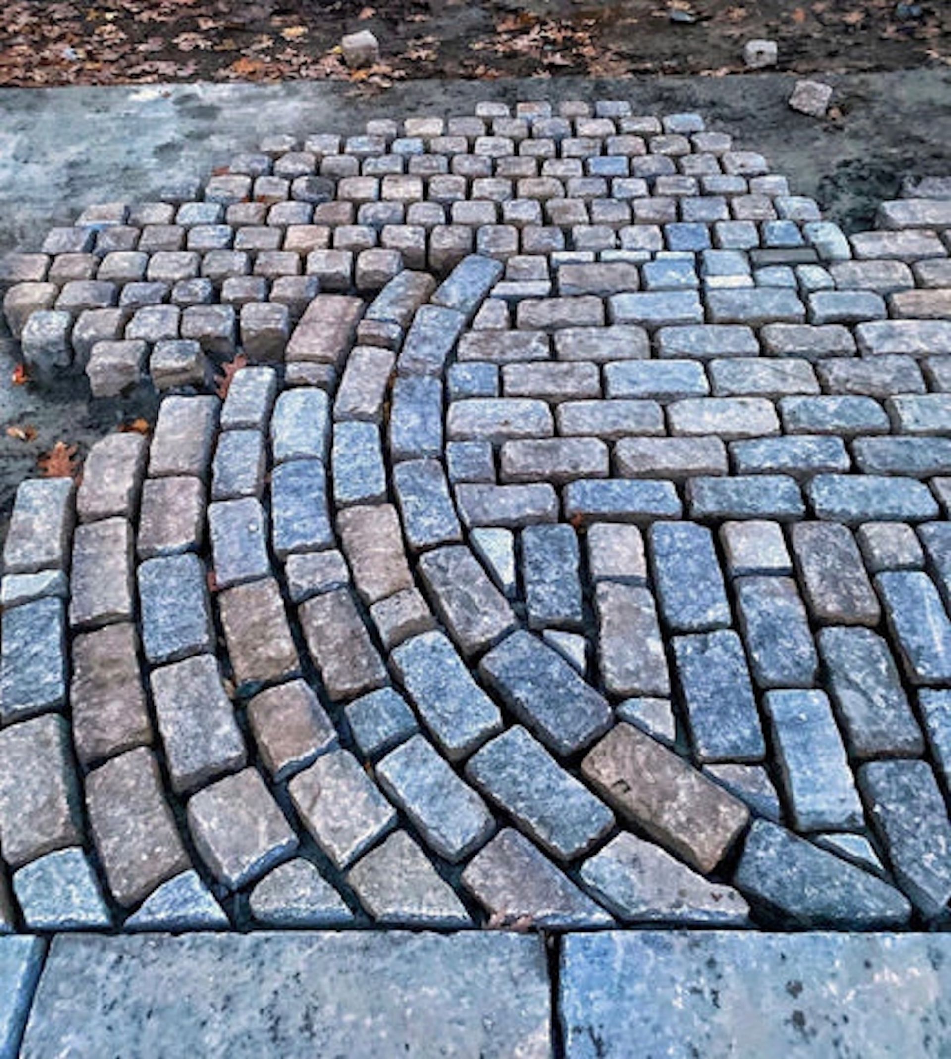 A close-up of a partially installed stone patio featuring a mix of grey and brown rectangular pavers in arched patterns.